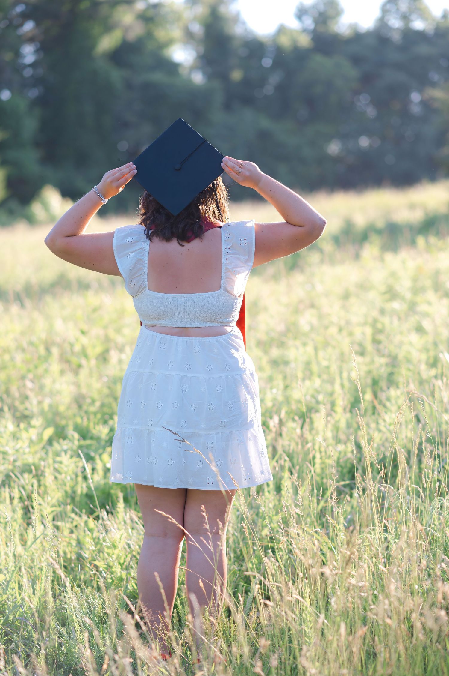 Back view of graduate in white dress holding cap while standing in golden field at sunset.
