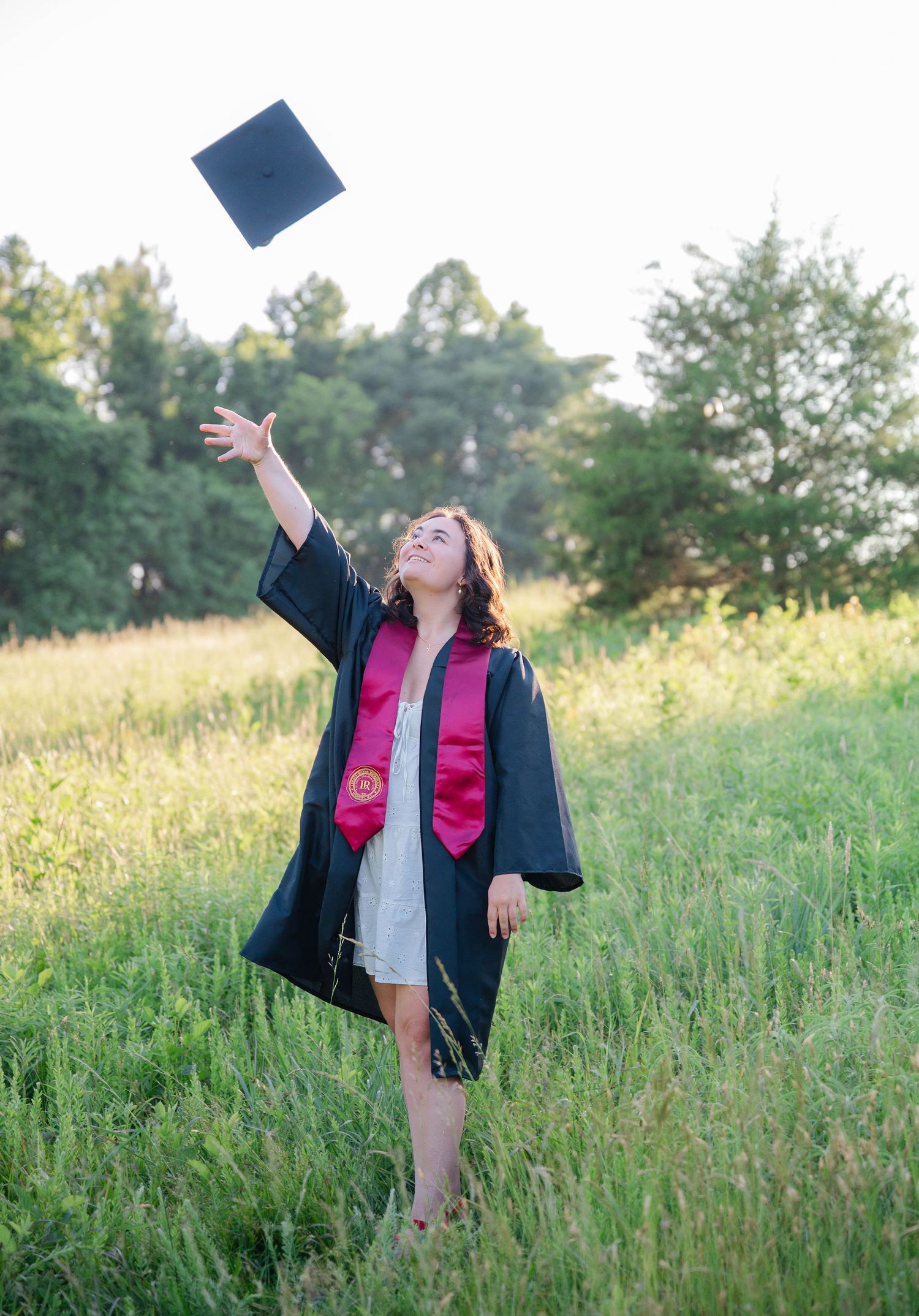 Graduate in cap and gown celebrates by tossing graduation cap in a sunny meadow.