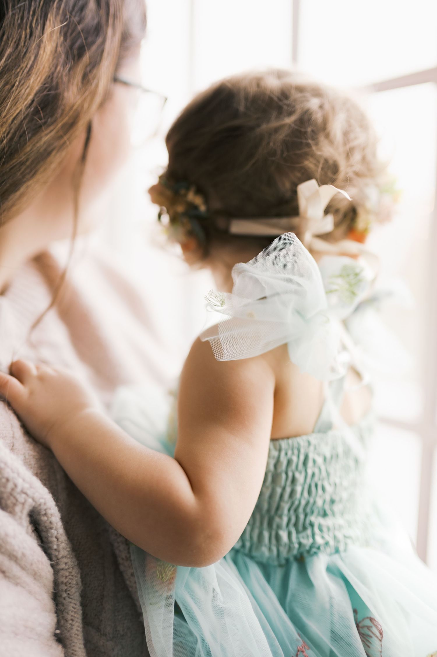 A young child in a mint green tulle dress looks out a bright window while standing beside someone.