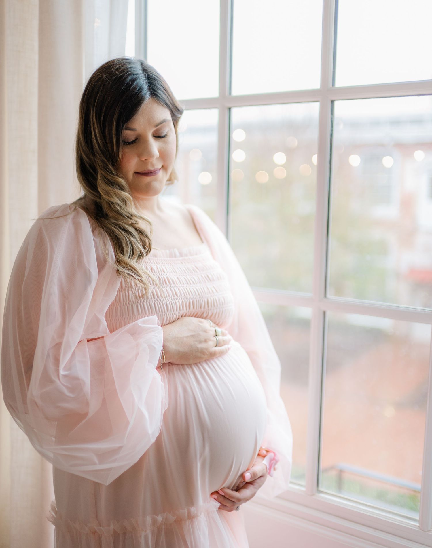 A maternity portrait by a window with soft natural light and bokeh in the background.