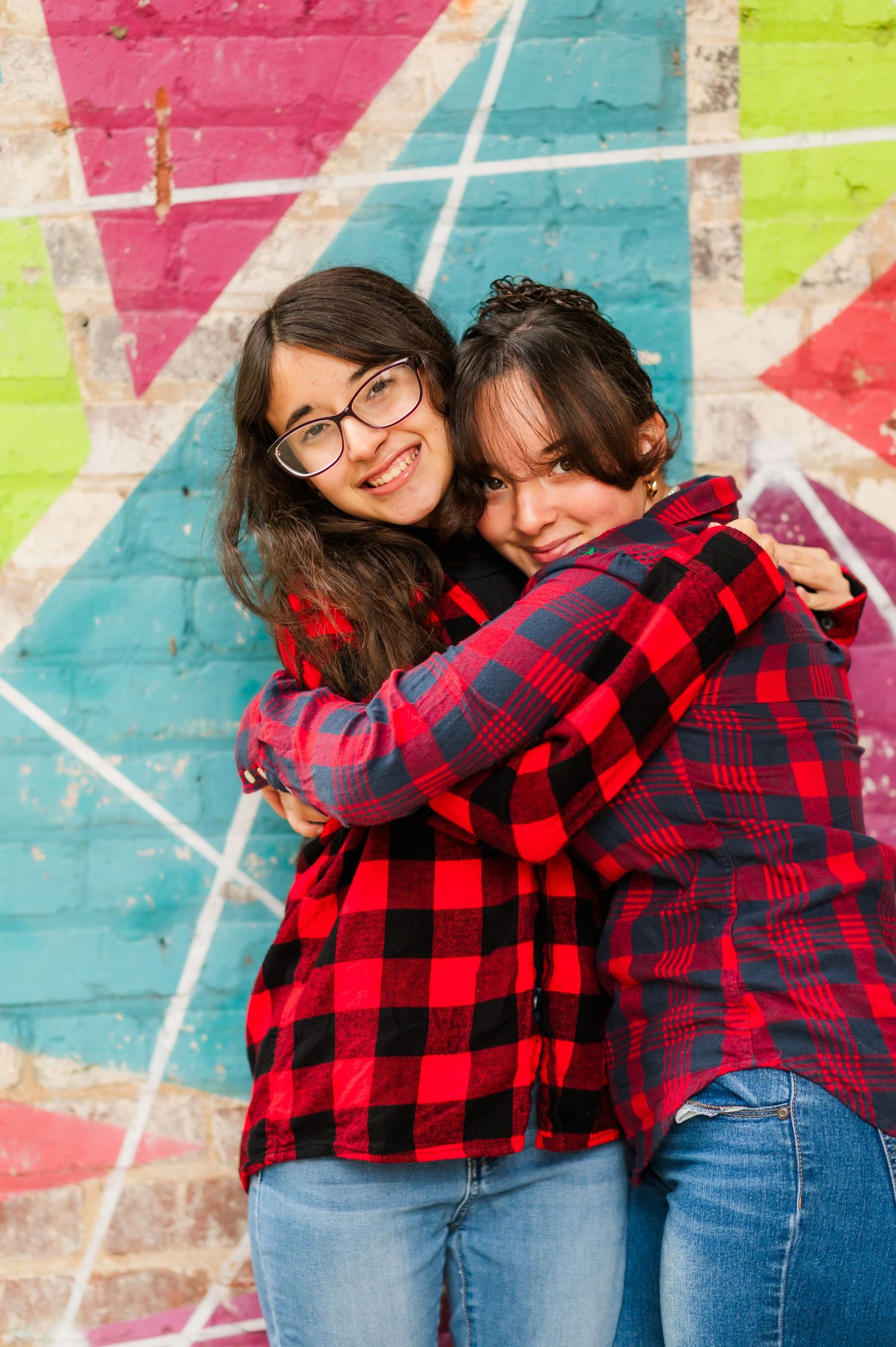 Friends in plaid shirts embrace while laughing against colorful geometric wall mural.