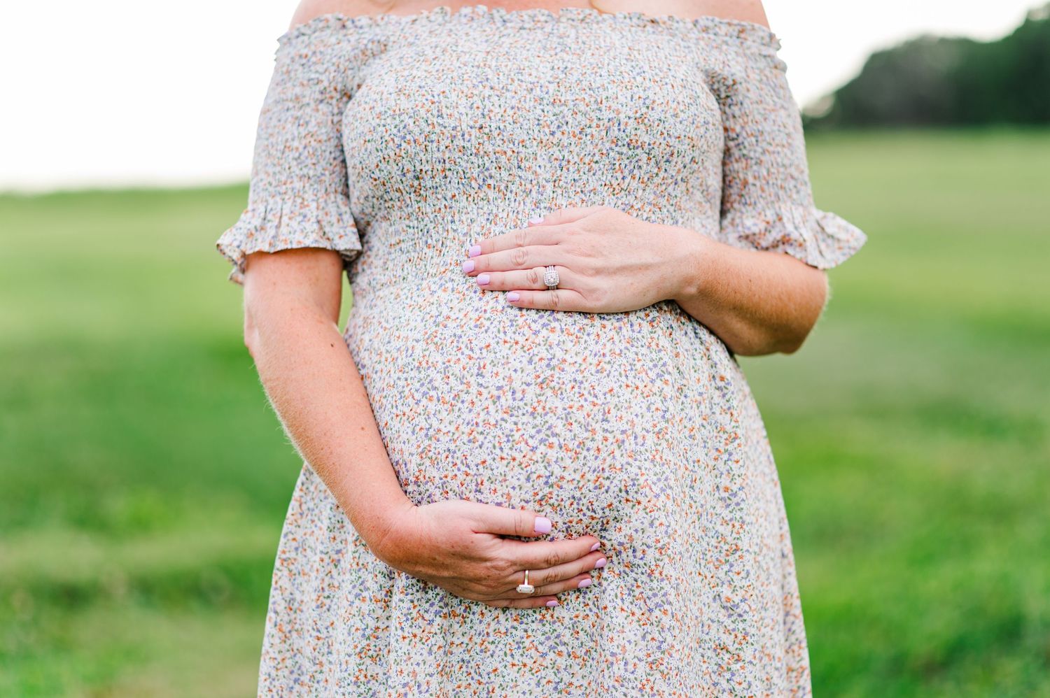 Close up of hands cradling a pregnant belly in a floral dress against a green outdoor background.