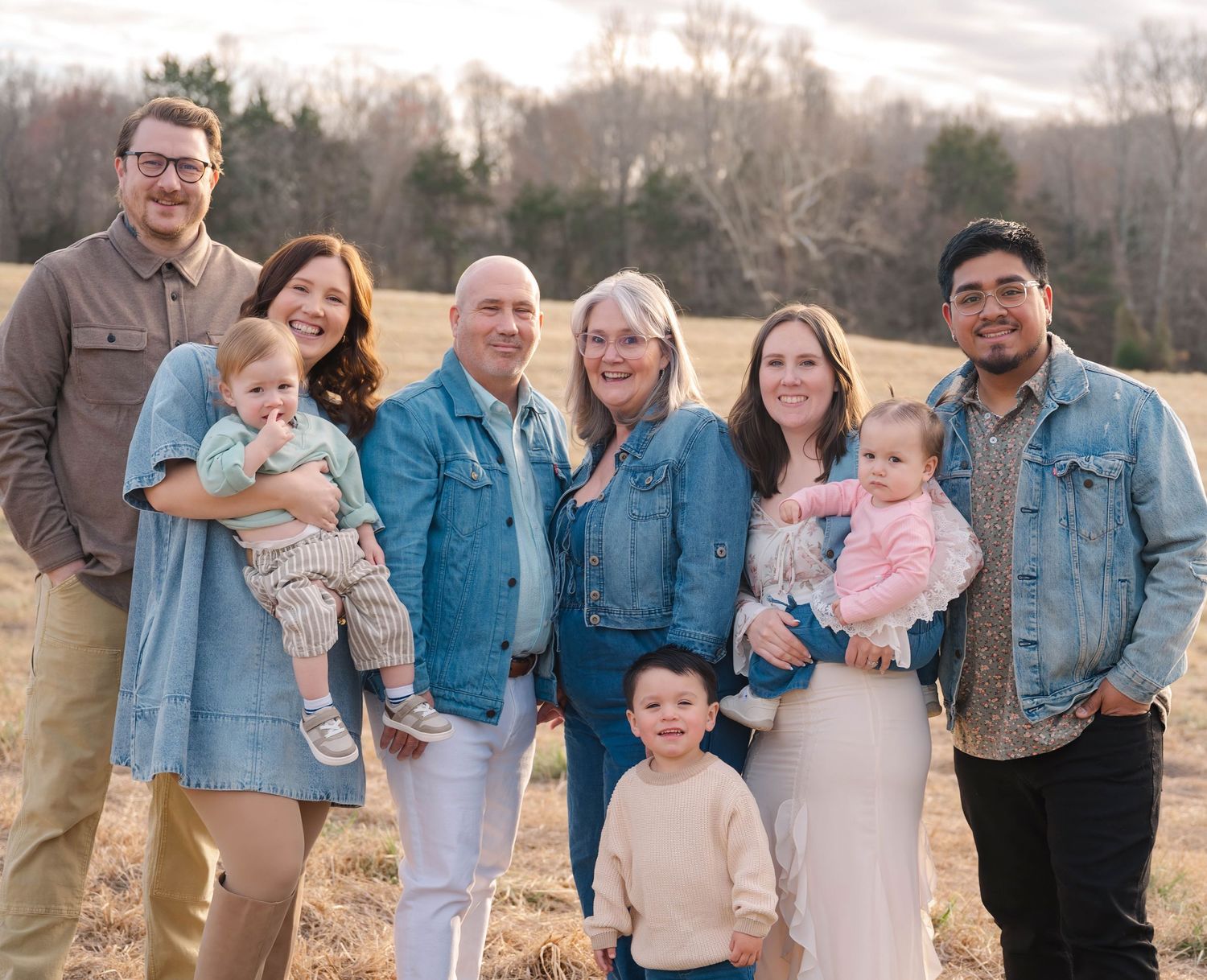 A group wearing coordinated denim jackets poses together in a sunny outdoor field during golden hour.