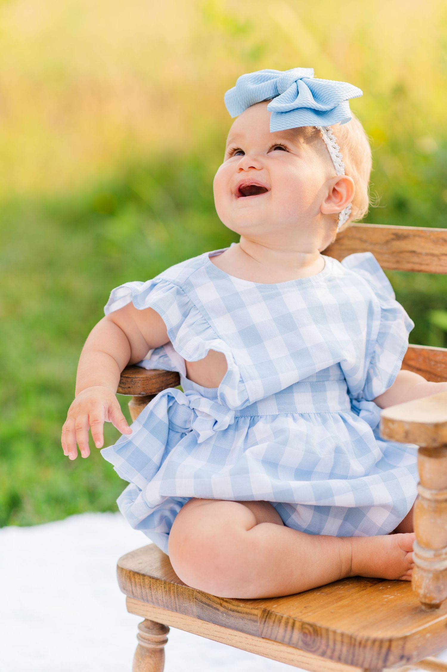 Baby wearing blue gingham dress sitting on wooden chair during outdoor portrait session.