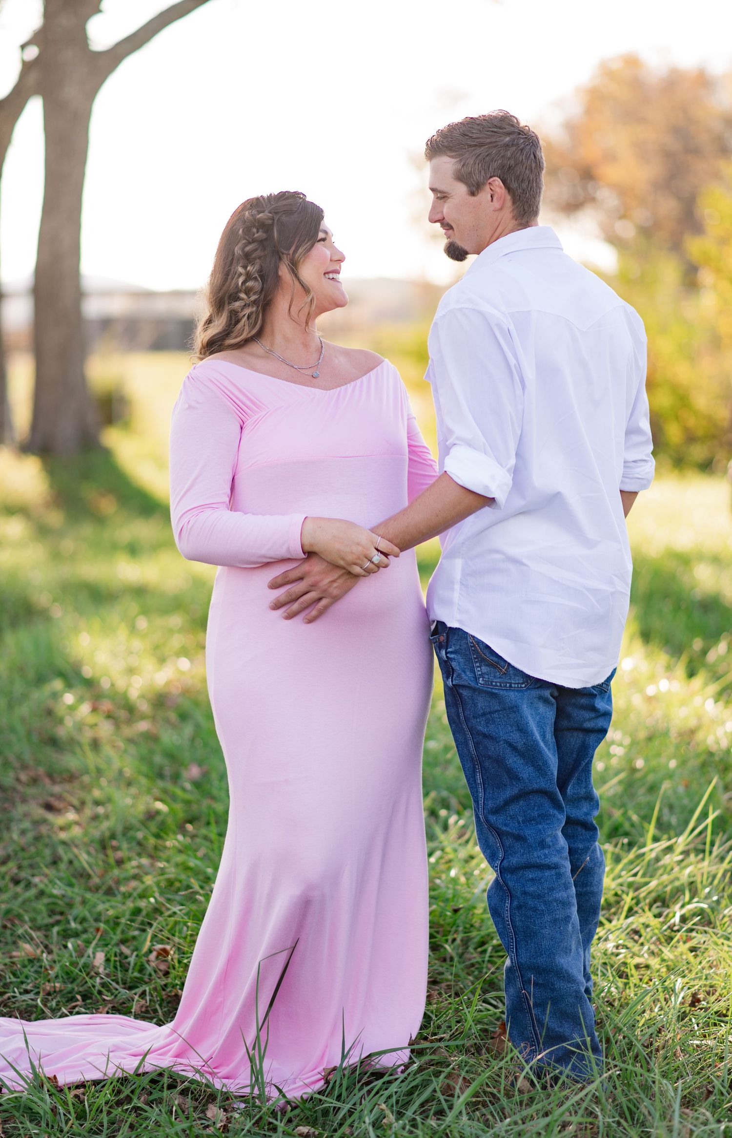 Couple in casual attire embracing during outdoor maternity session in grassy field at sunset.