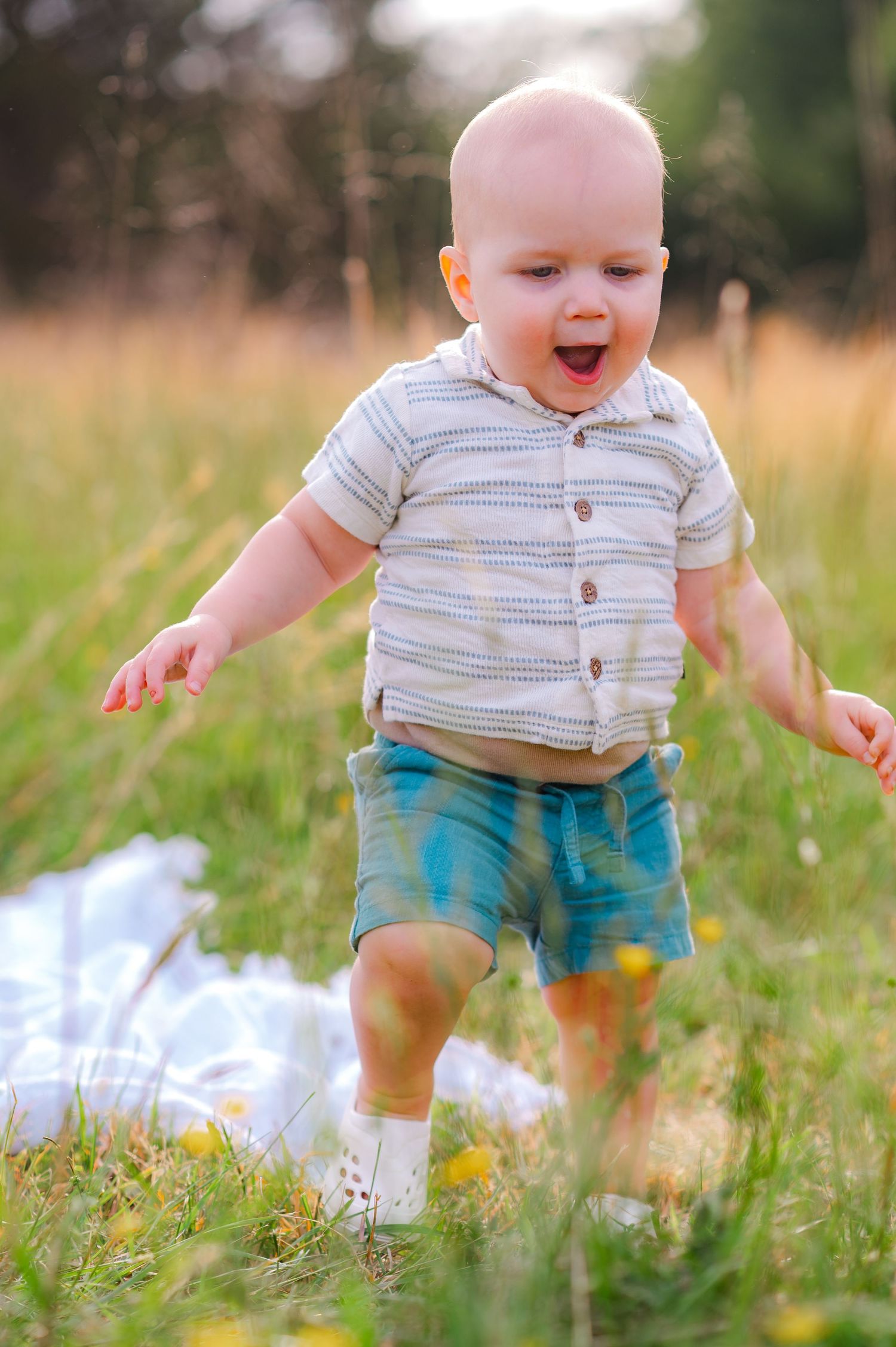 Baby in striped shirt and blue shorts playing in grassy field during summer.