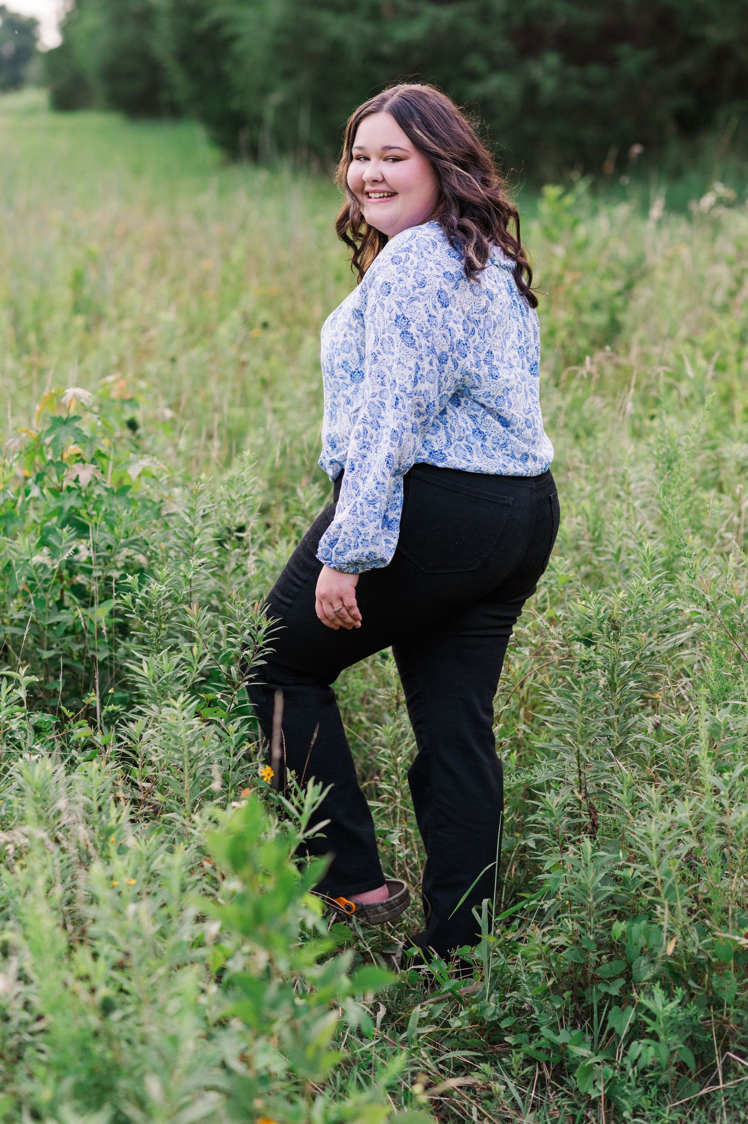Woman in floral top and black pants poses playfully in a green meadow.