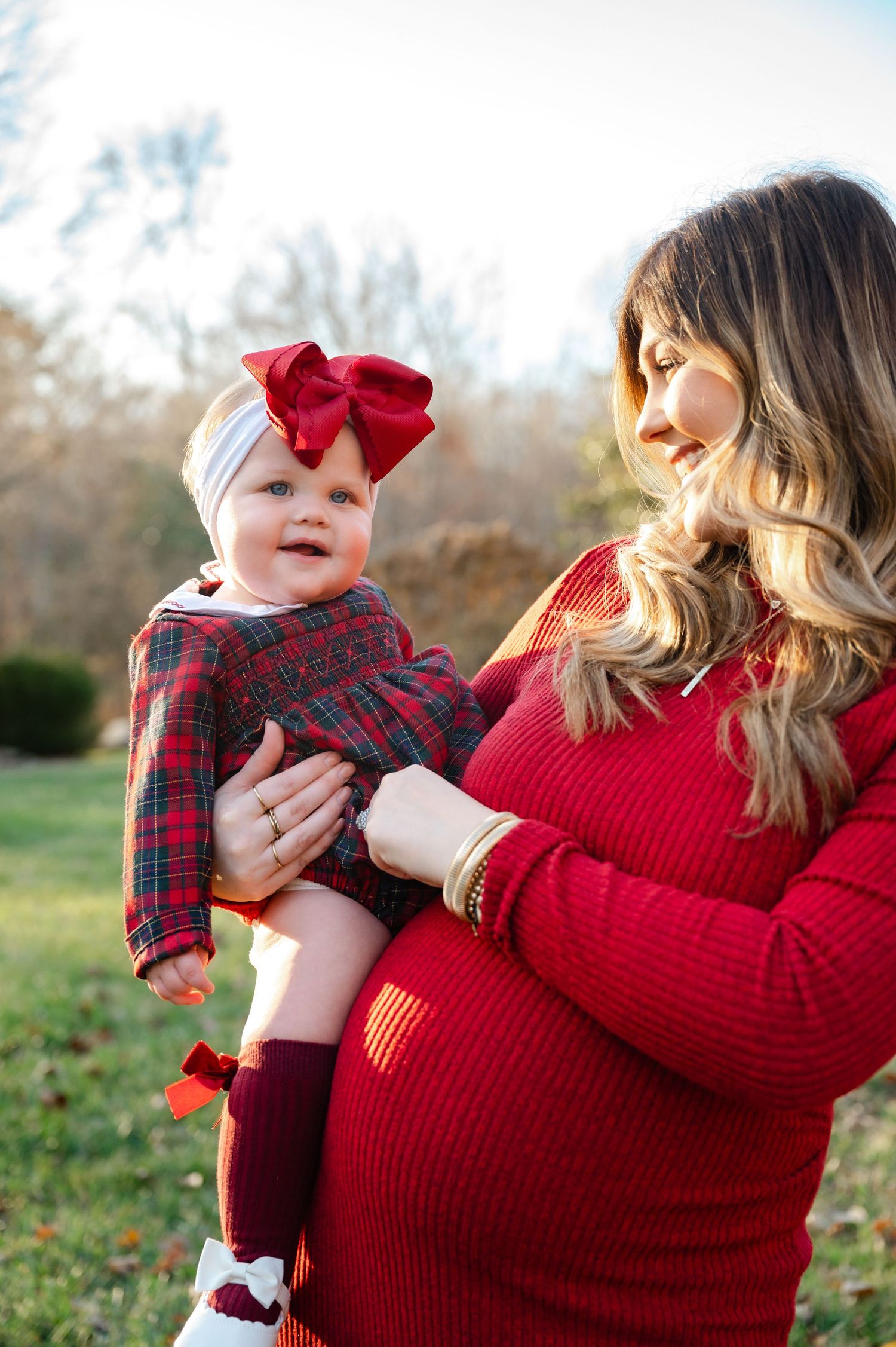 Mother in red sweater holds baby wearing festive plaid outfit and bow in outdoor setting.