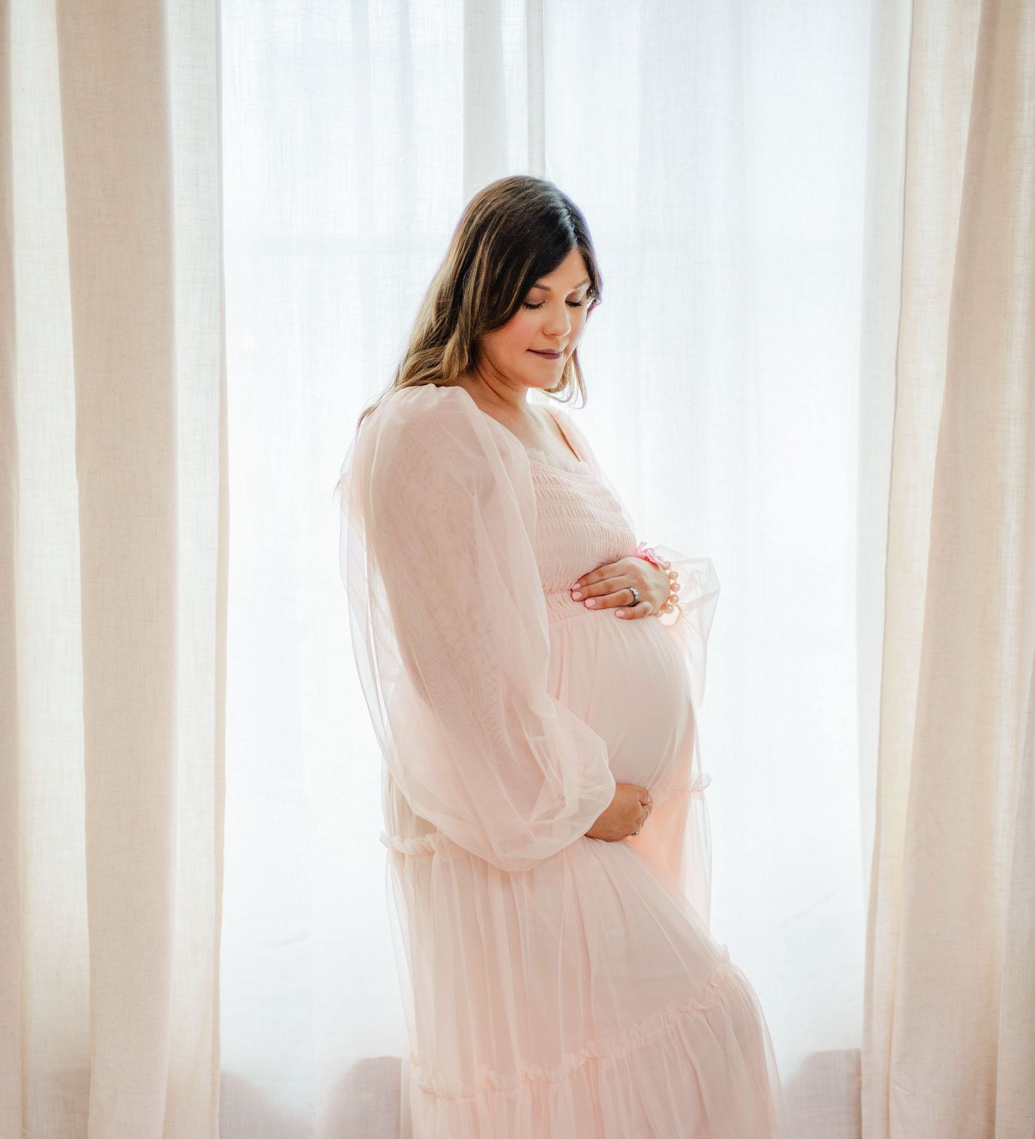 Ethereal maternity portraits in flowing pink dress against white curtained window.