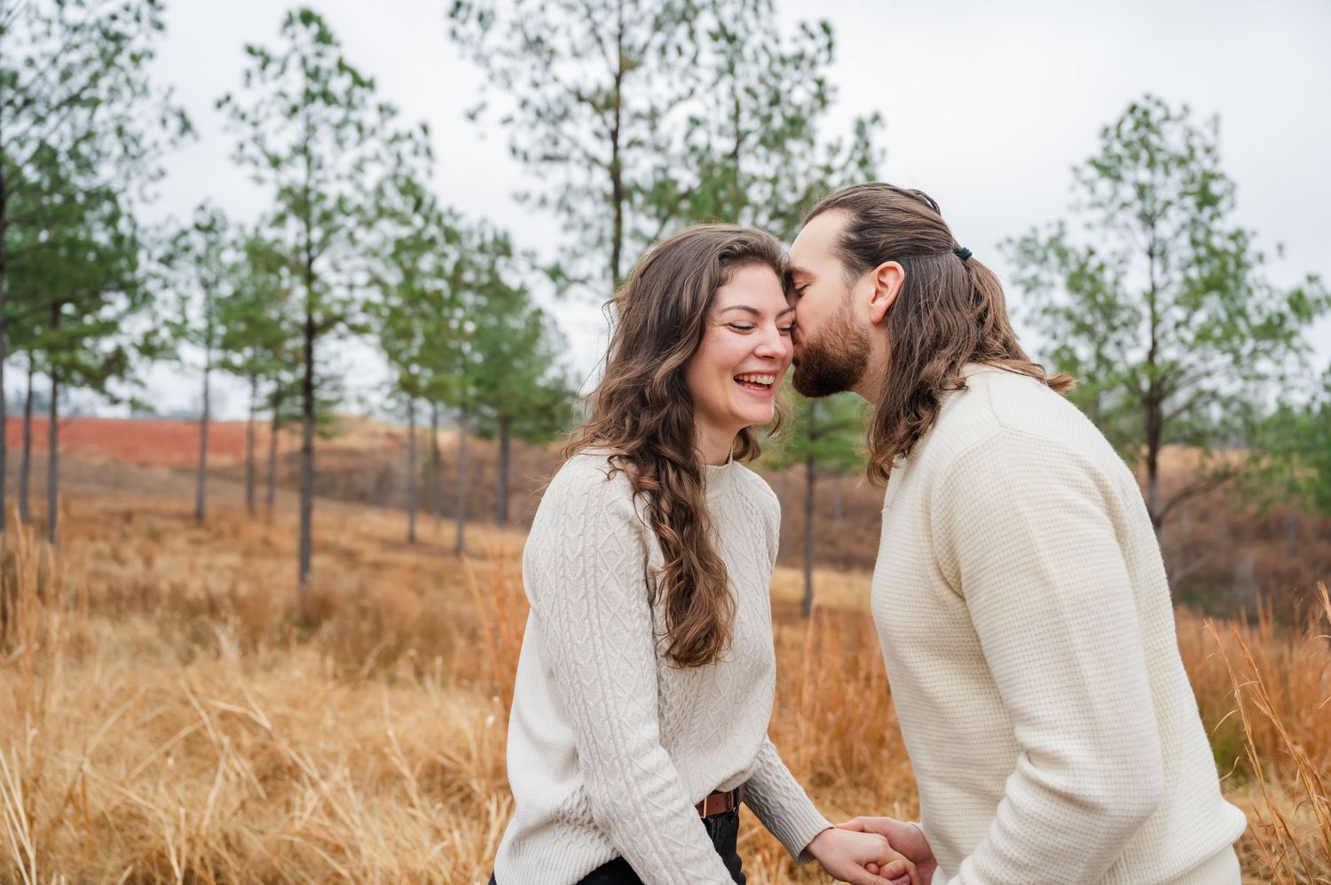 A romantic couple in cream sweaters share a joyful moment together in a rustic outdoor field setting.