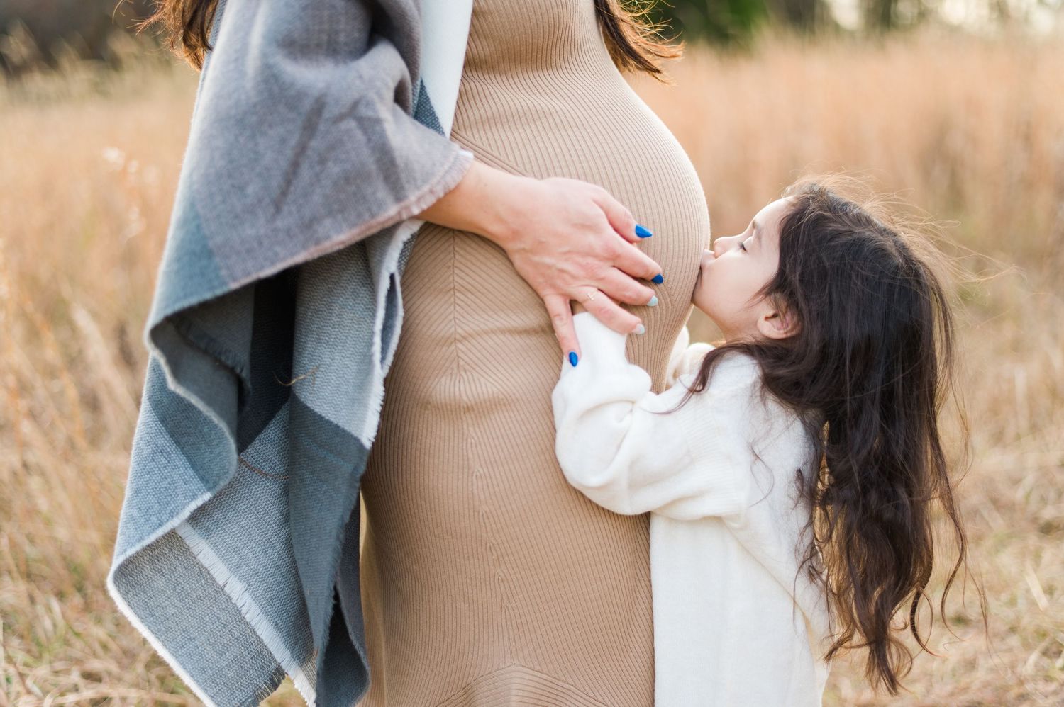 Woman in beige dress and gray cardigan poses outdoors during pregnancy photoshoot.