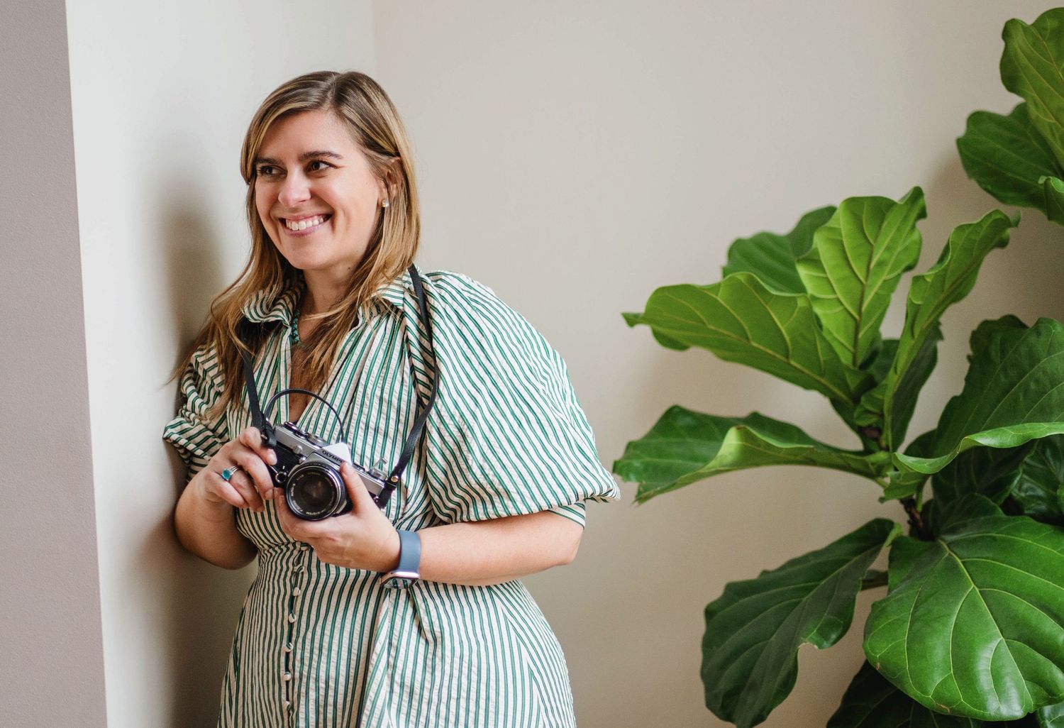 A photographer in a striped shirt holds a vintage camera next to a large fiddle leaf fig plant.