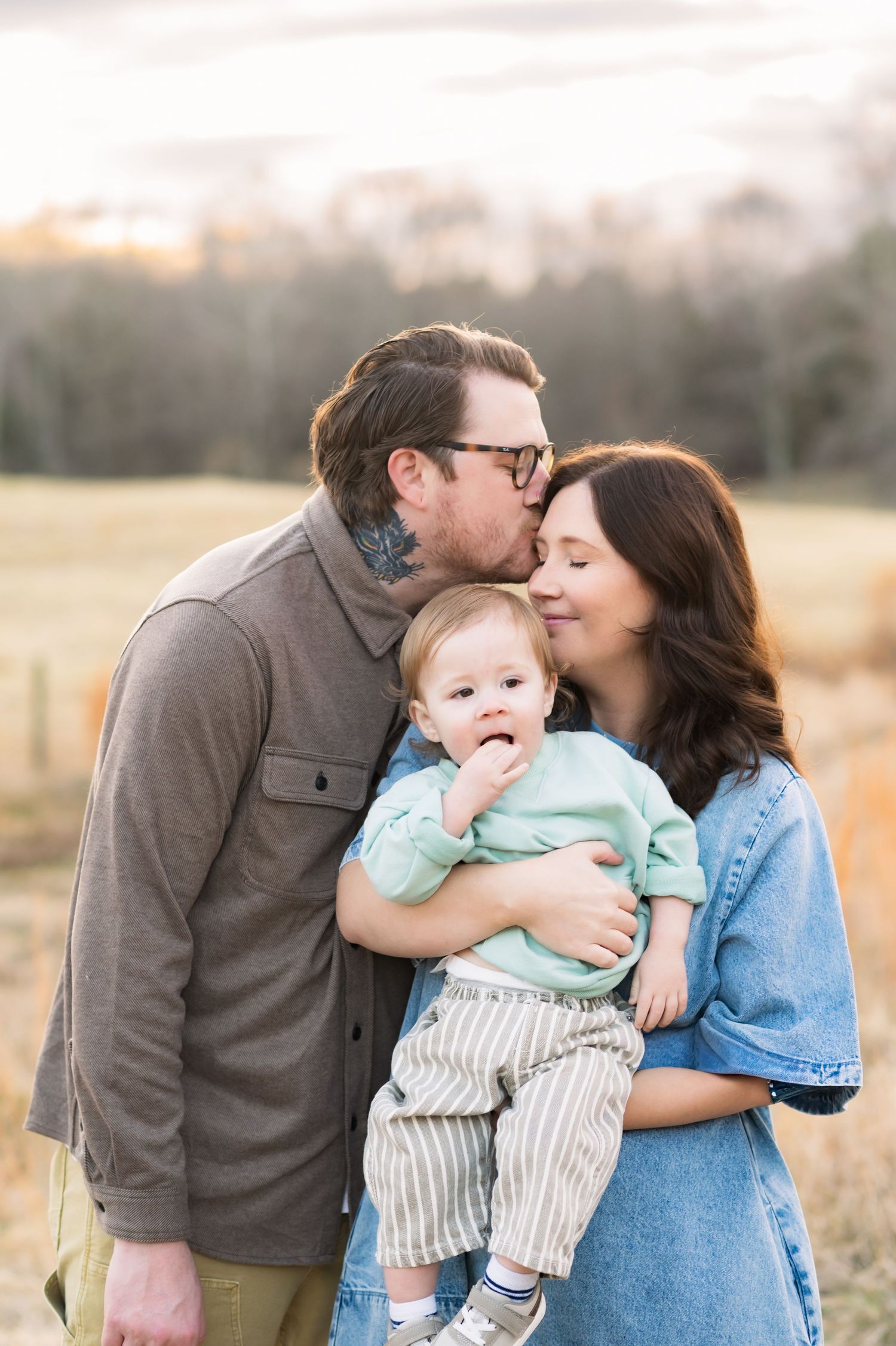 A tender family moment in a sunlit field as parents share affection while holding their baby in casual autumn attire.