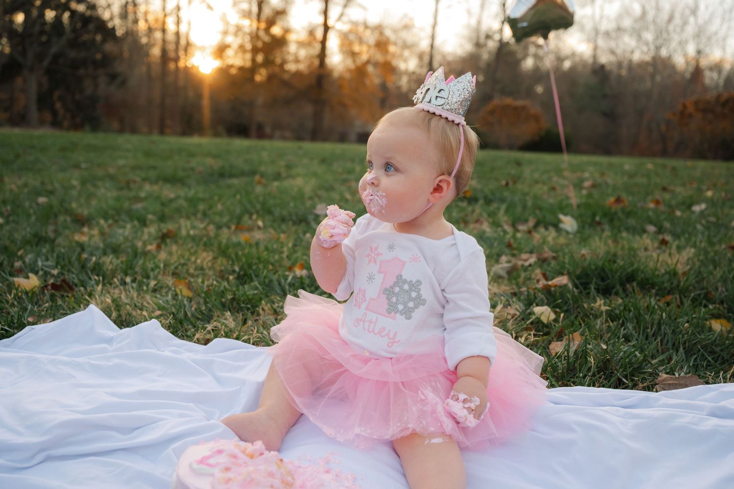 Baby in pink tutu dress sitting on white blanket outdoors during sunset photoshoot.