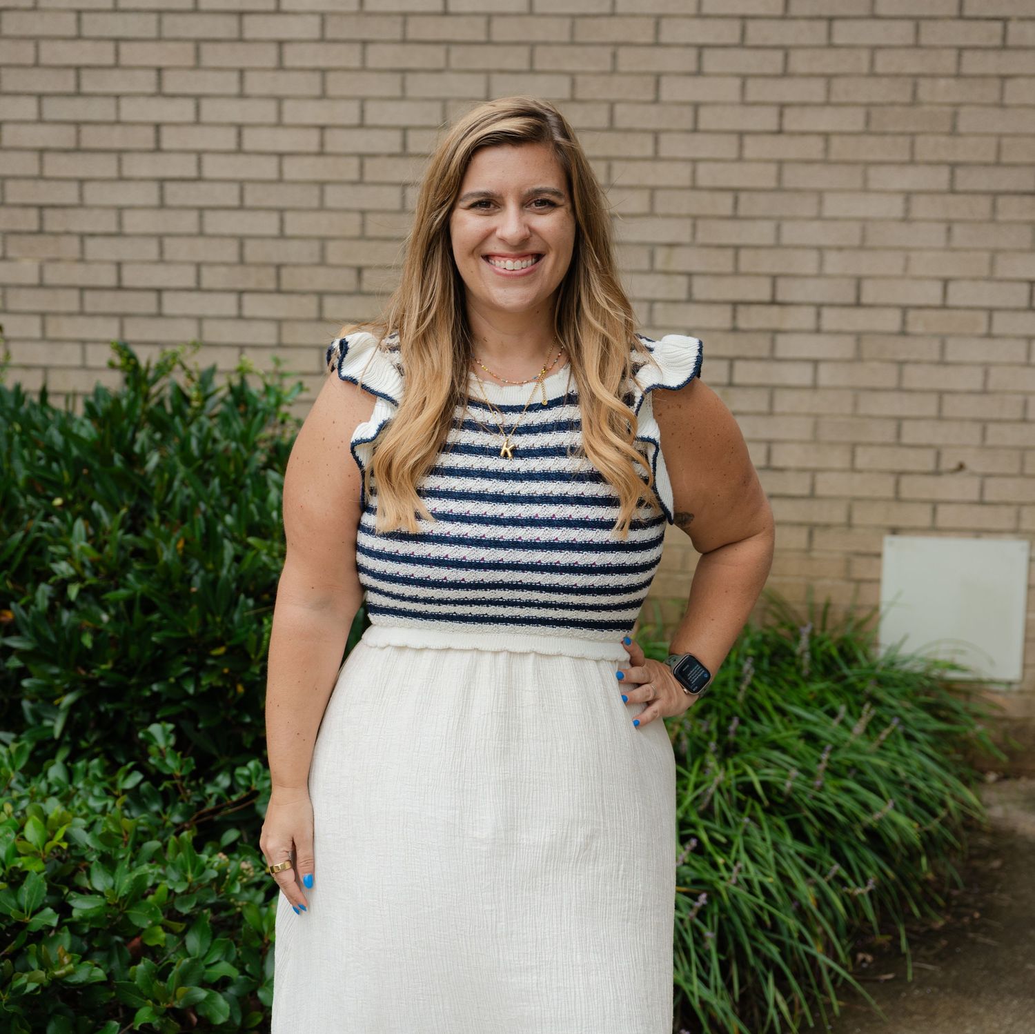 Woman in a navy striped and white dress poses against a brick wall with greenery in the background.