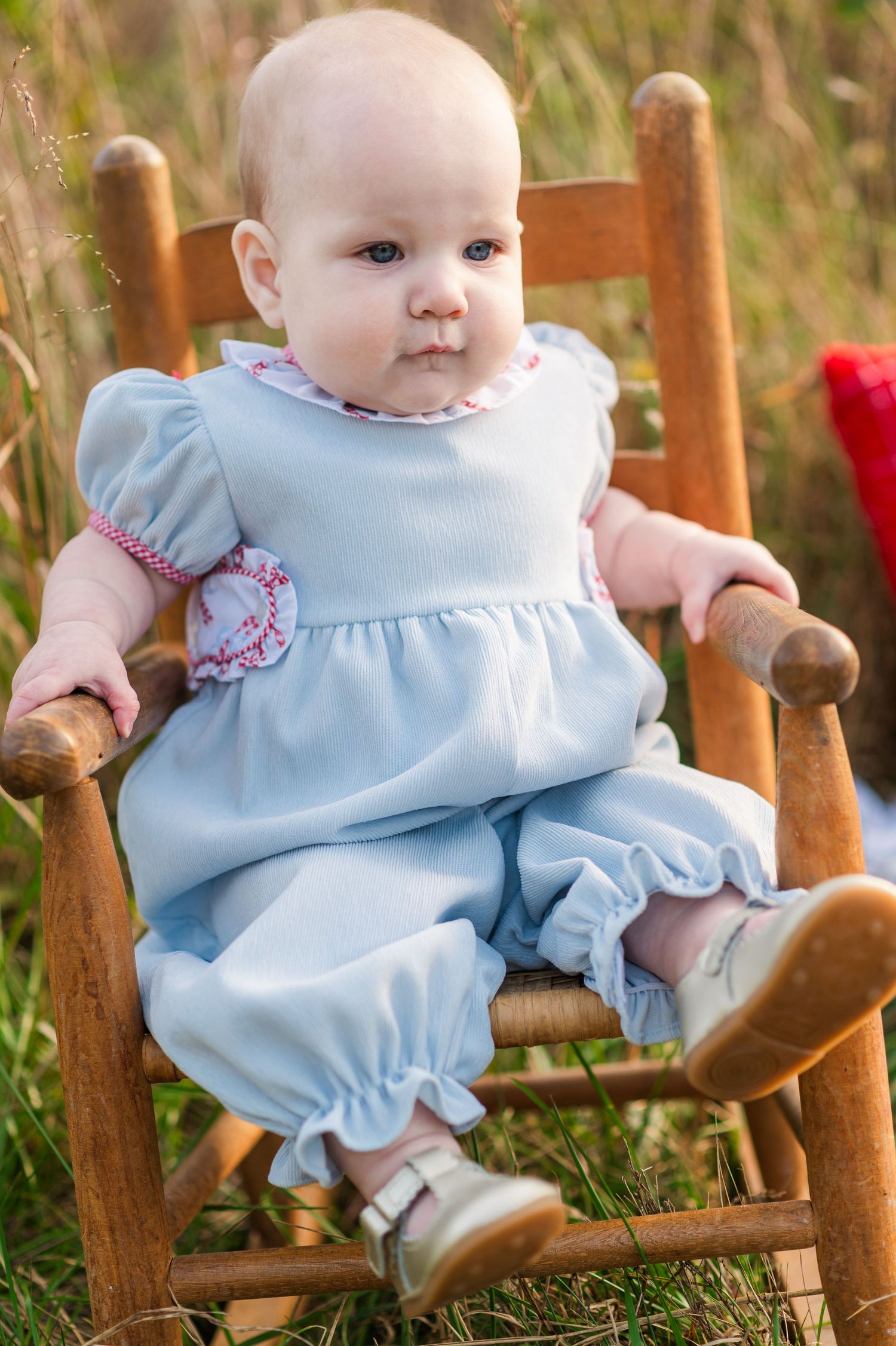 Baby in light blue romper sitting in wooden rocking chair outdoors among tall grass.