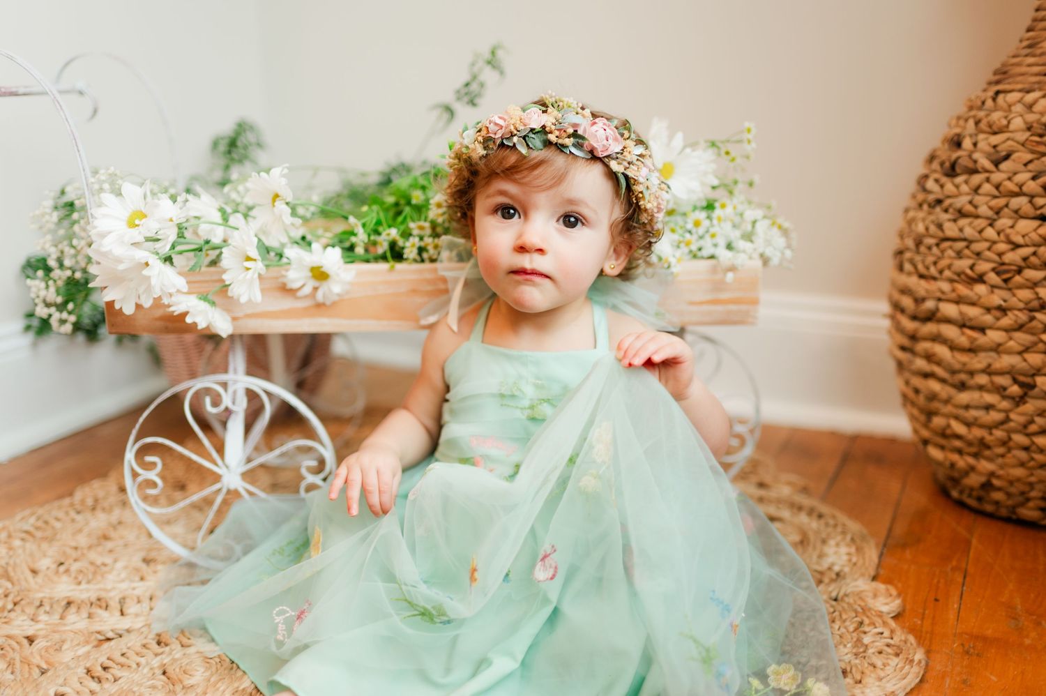 A toddler in a mint green tulle dress and floral crown sits on a hardwood floor near white flowers and decor.