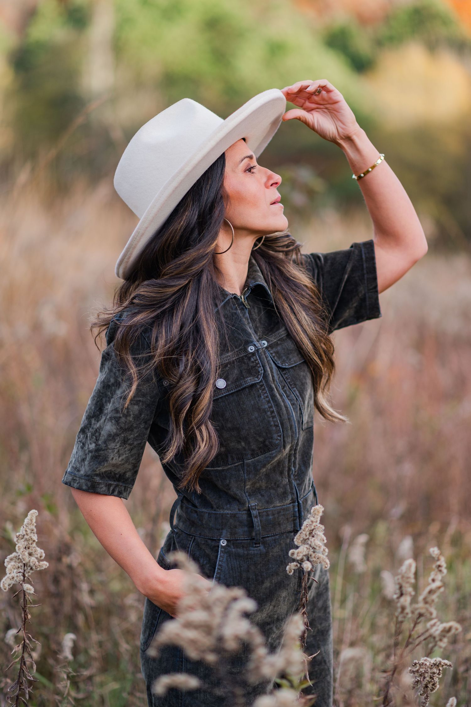Woman in denim outfit and white hat poses dramatically in field during golden hour.