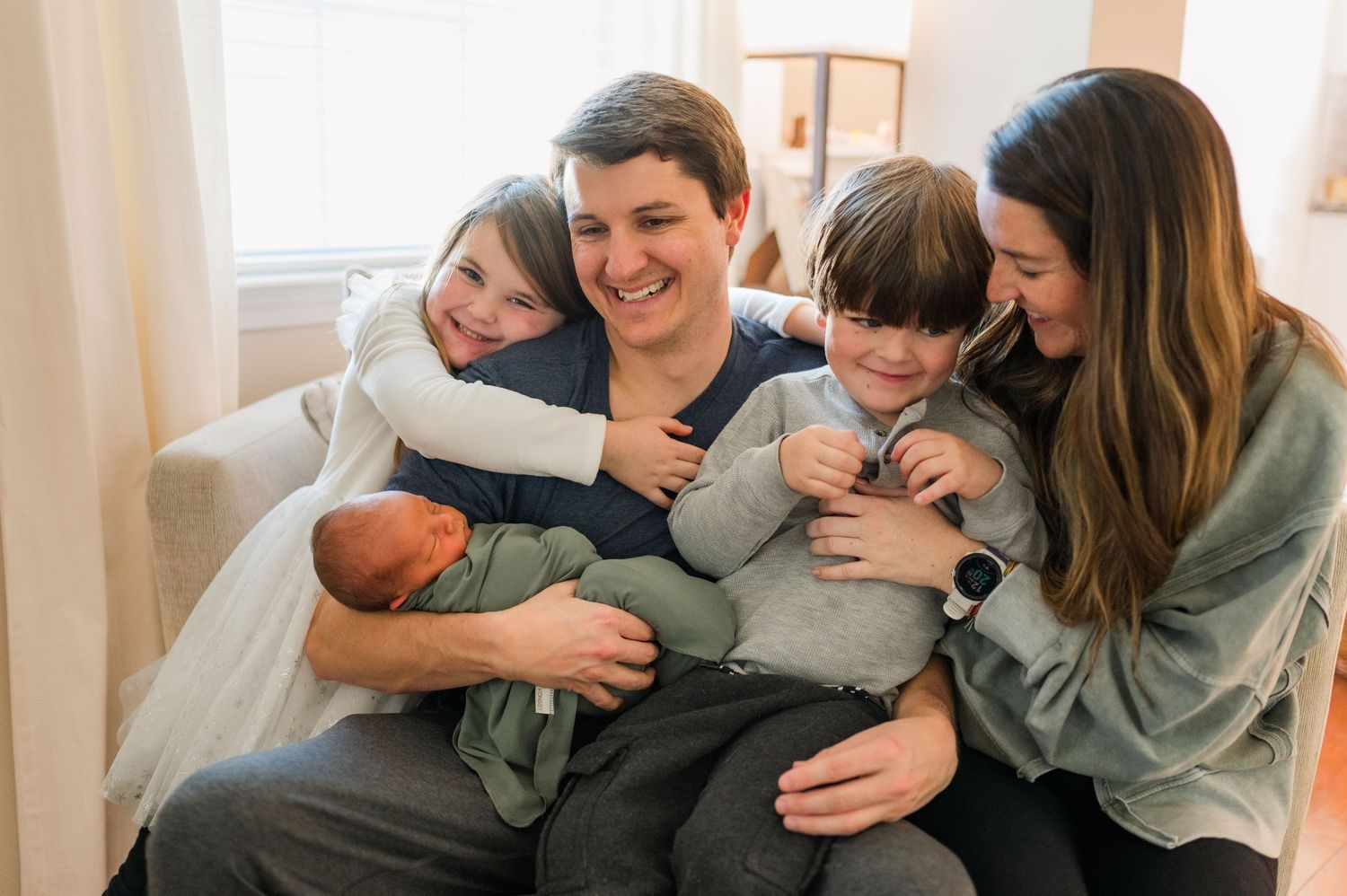 A family wearing matching grey sweaters embraces and laughs together in a bright room with white curtains.