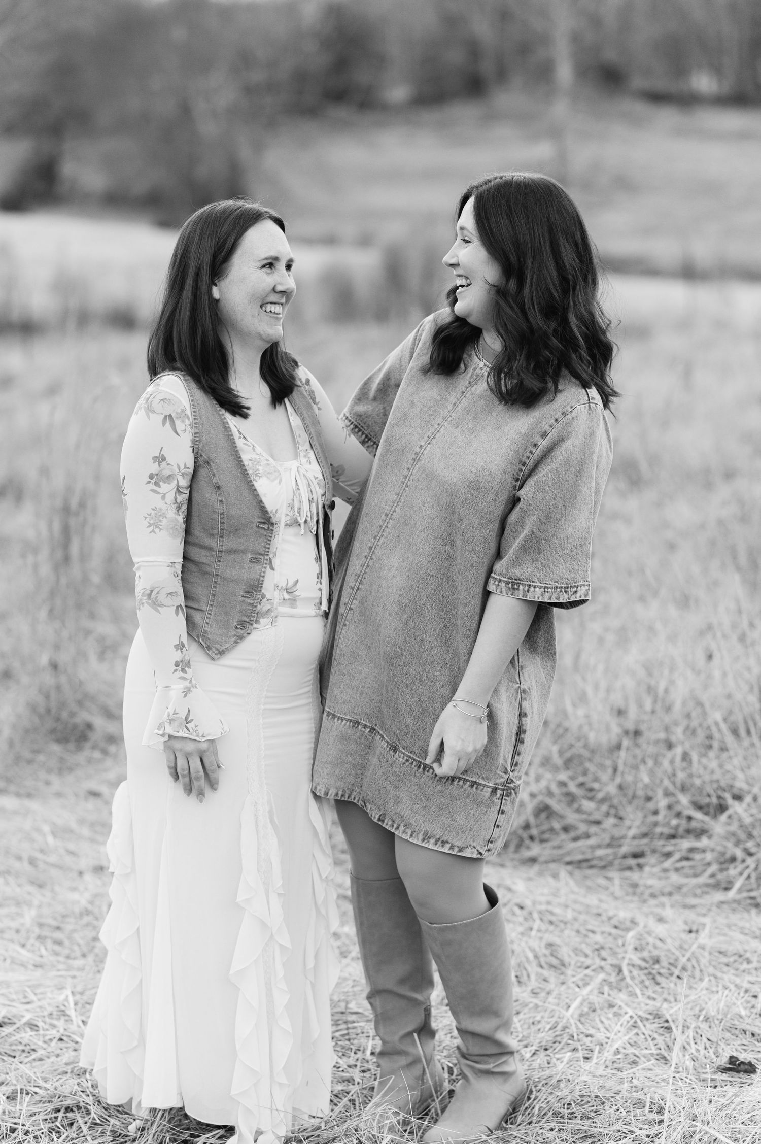 Two women share a joyful laugh together in a field, one wearing a white dress and vest, the other in denim.