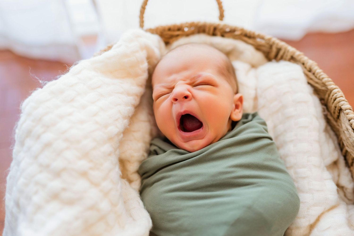A newborn baby yawning while wrapped in olive green swaddle in a woven basket.