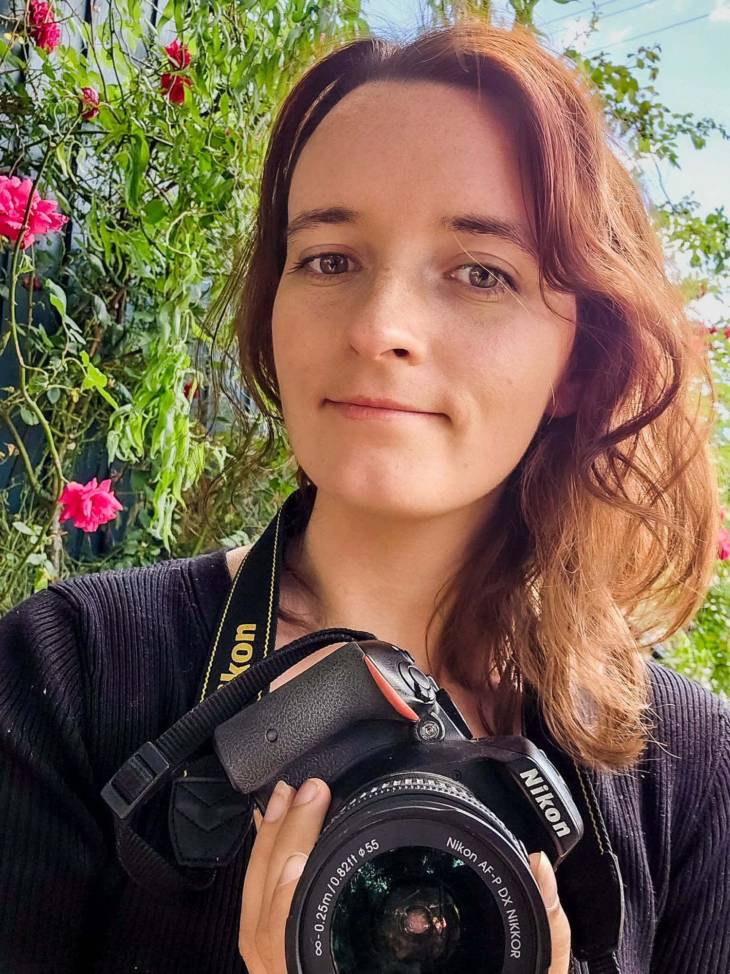 Photographer with curly hair holding professional camera against floral background.