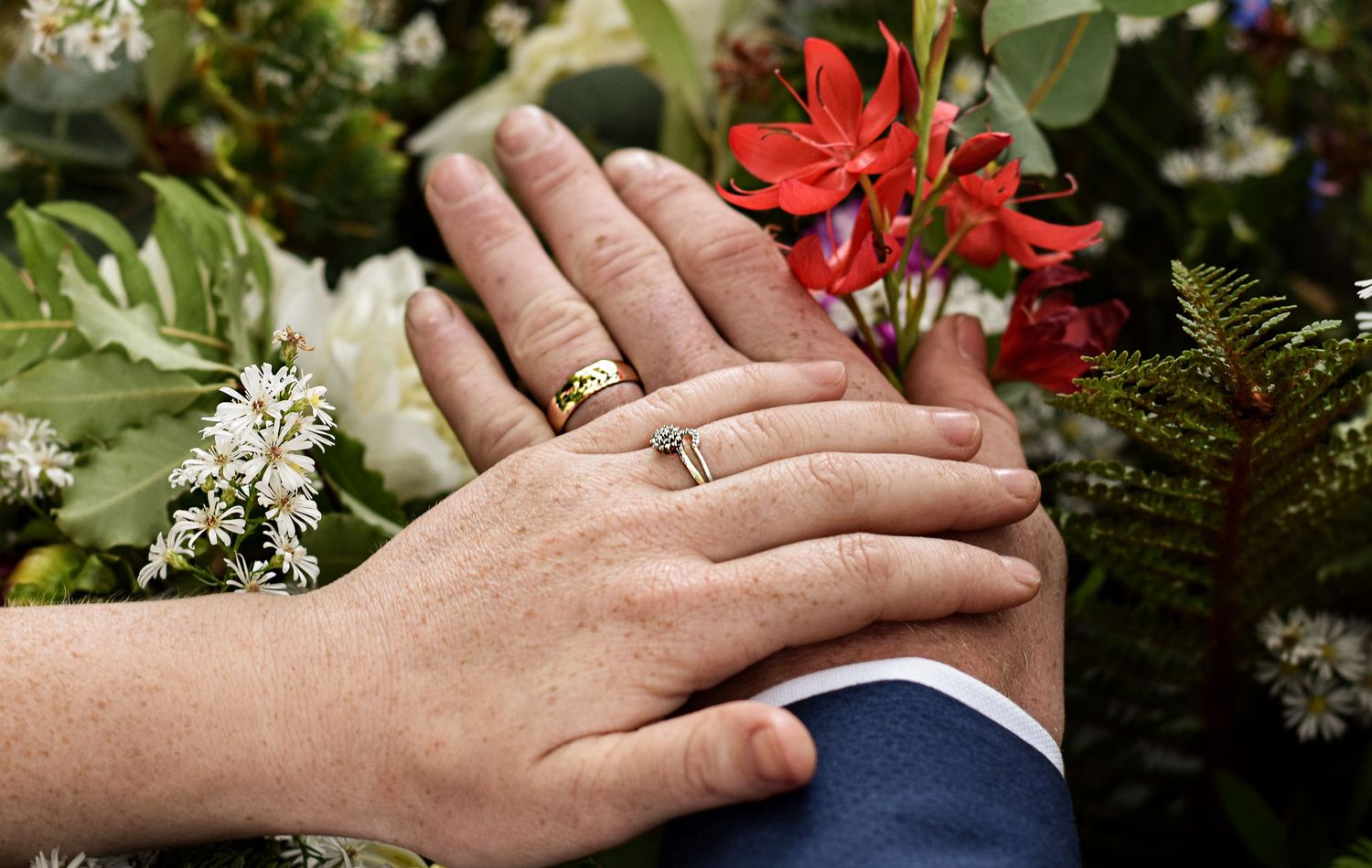 Close-up of hands with wedding rings resting on formal attire with red and white flowers.