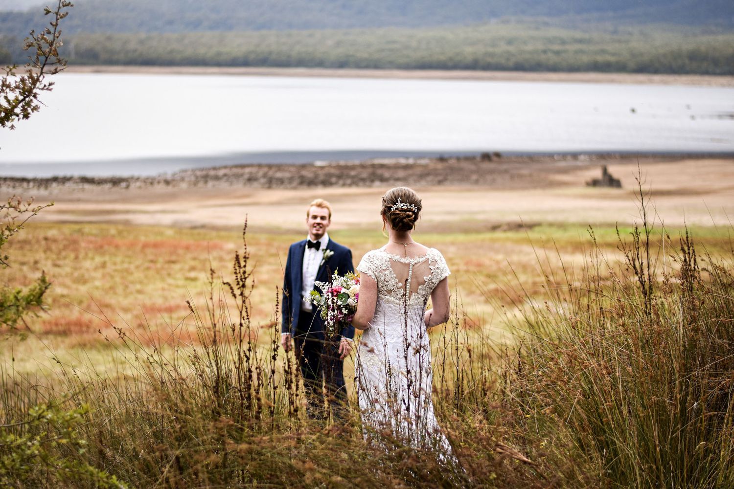 Newlyweds walking through scenic mountain meadow with lake view.