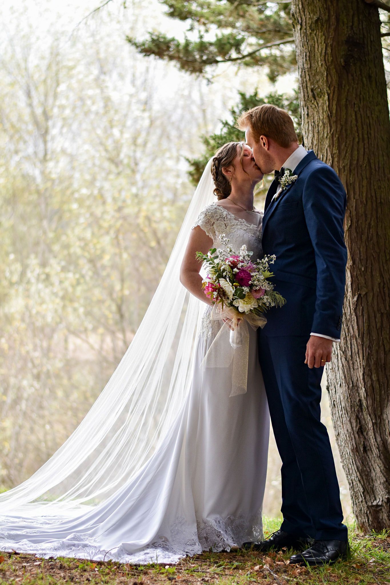 A romantic wedding moment captured between a couple in white dress and navy suit beneath tall trees.