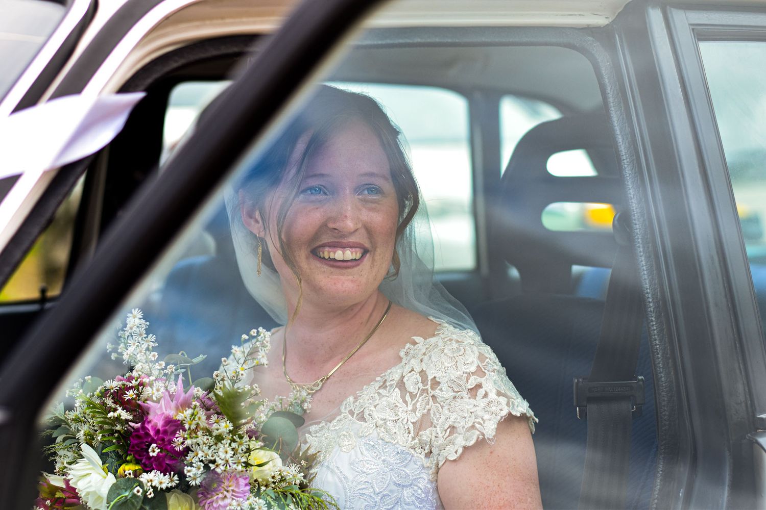 Person in bridal attire smiling while seated in car with floral bouquet.