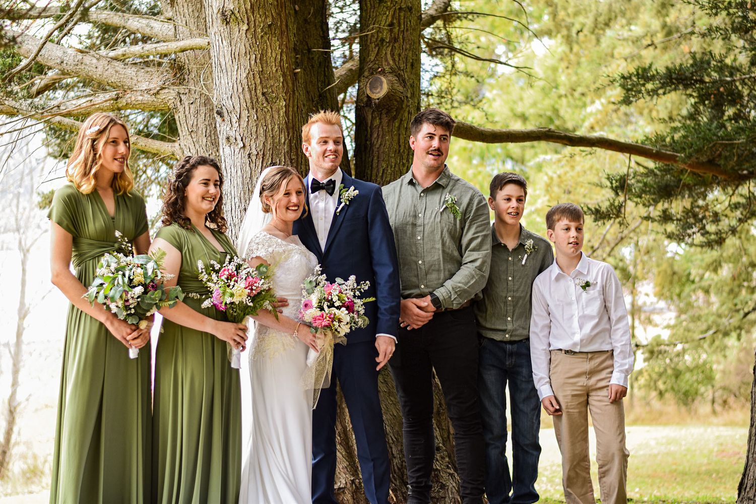 Wedding party in light green dresses poses together outdoors under tall pine trees.