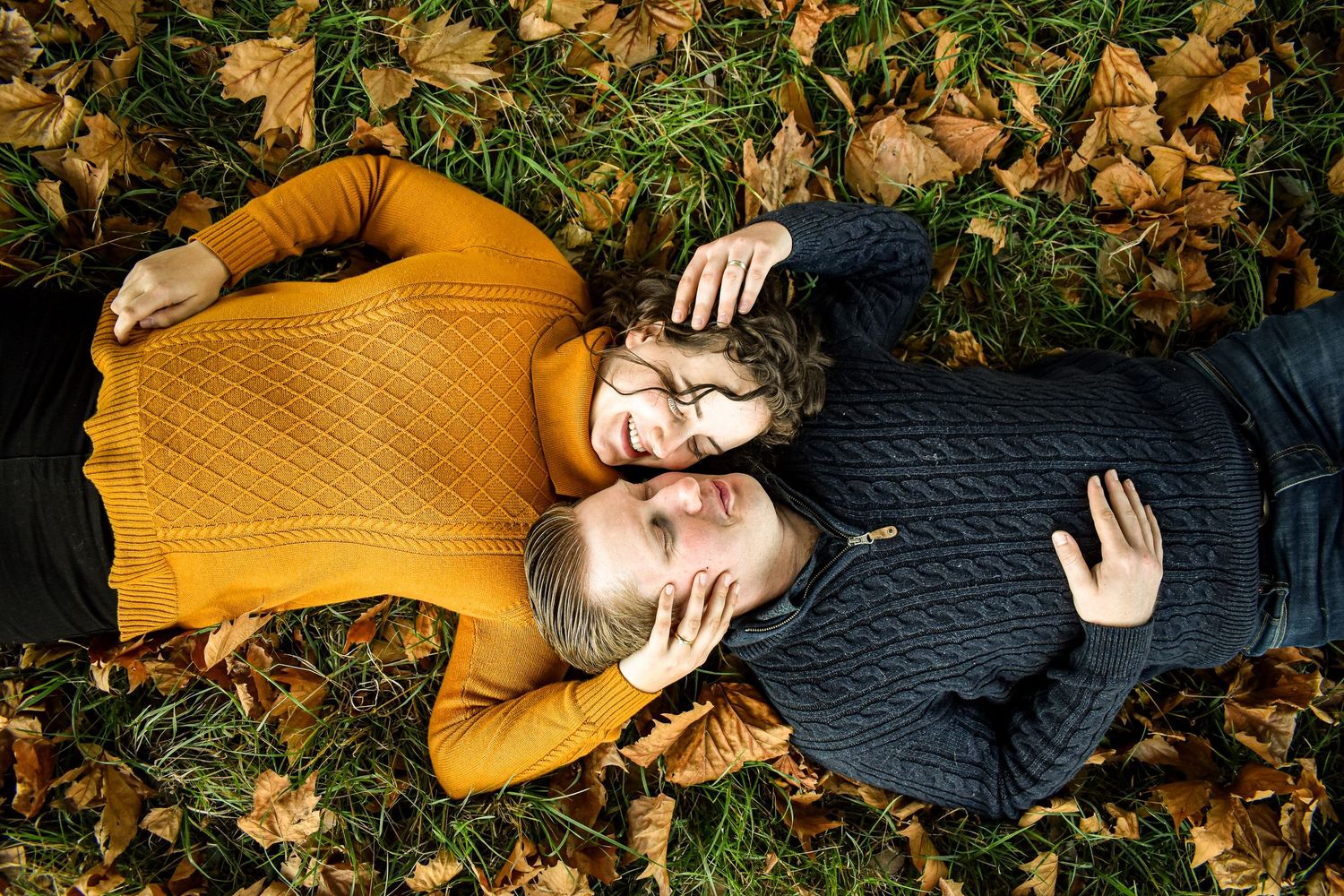 Two people lying in autumn leaves wearing mustard yellow and navy sweaters during a fall photoshoot.