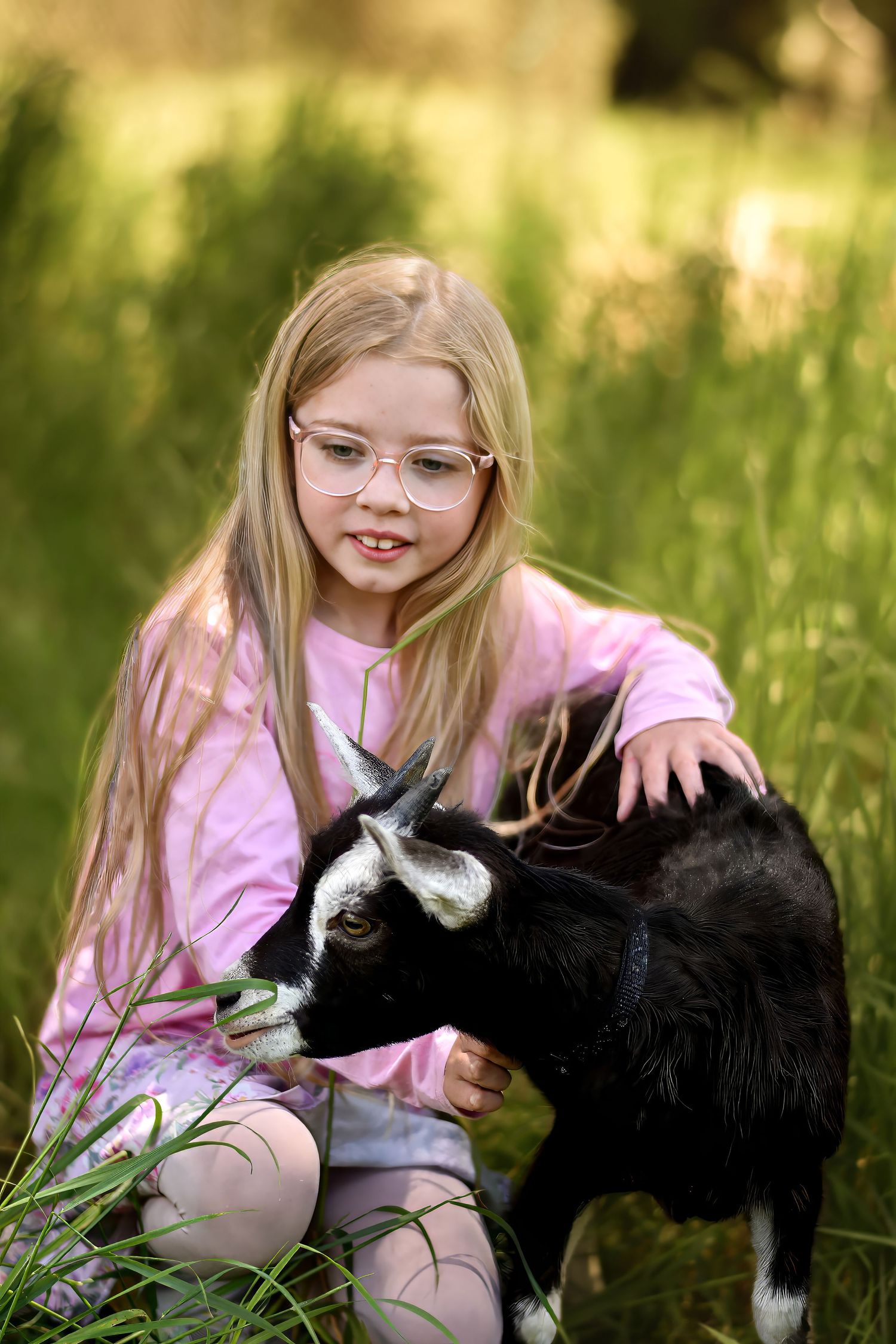 Young person in pink shirt petting a black and white goat in a sunny meadow.