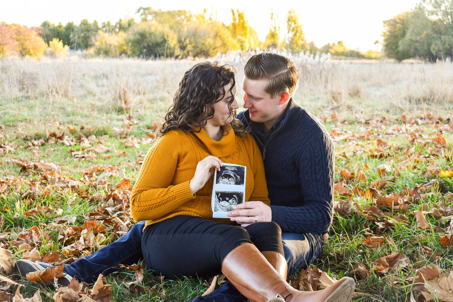 Couple sitting on autumn leaves holding ultrasound photo during outdoor photoshoot.