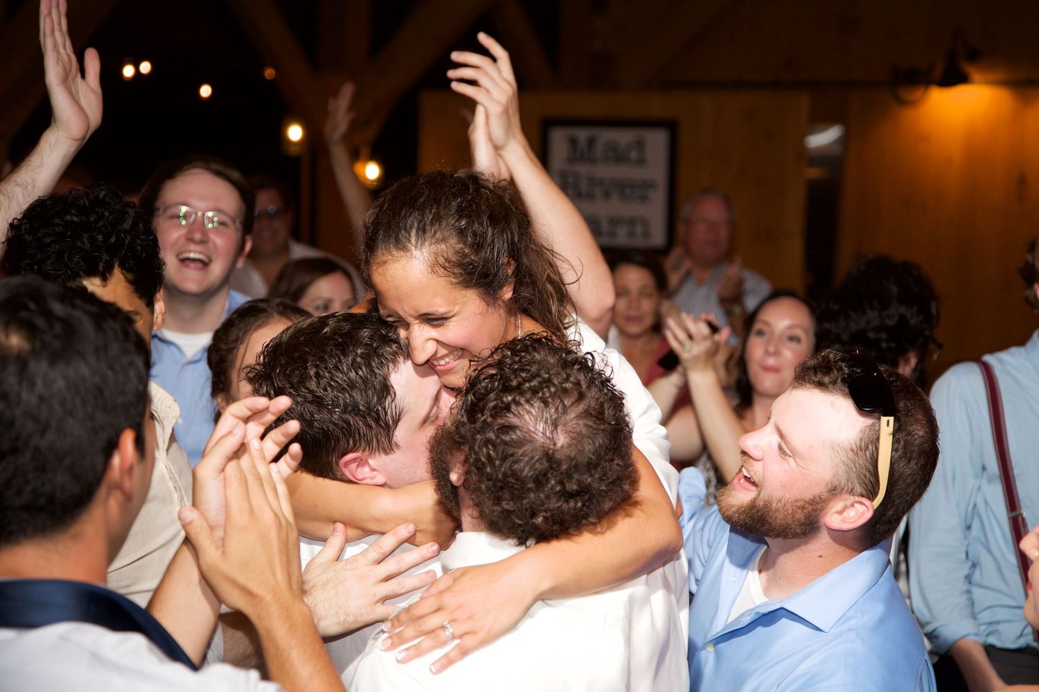 Joyful wedding reception crowd lifting the bride and groom in a lively celebration inside a rustic venue.