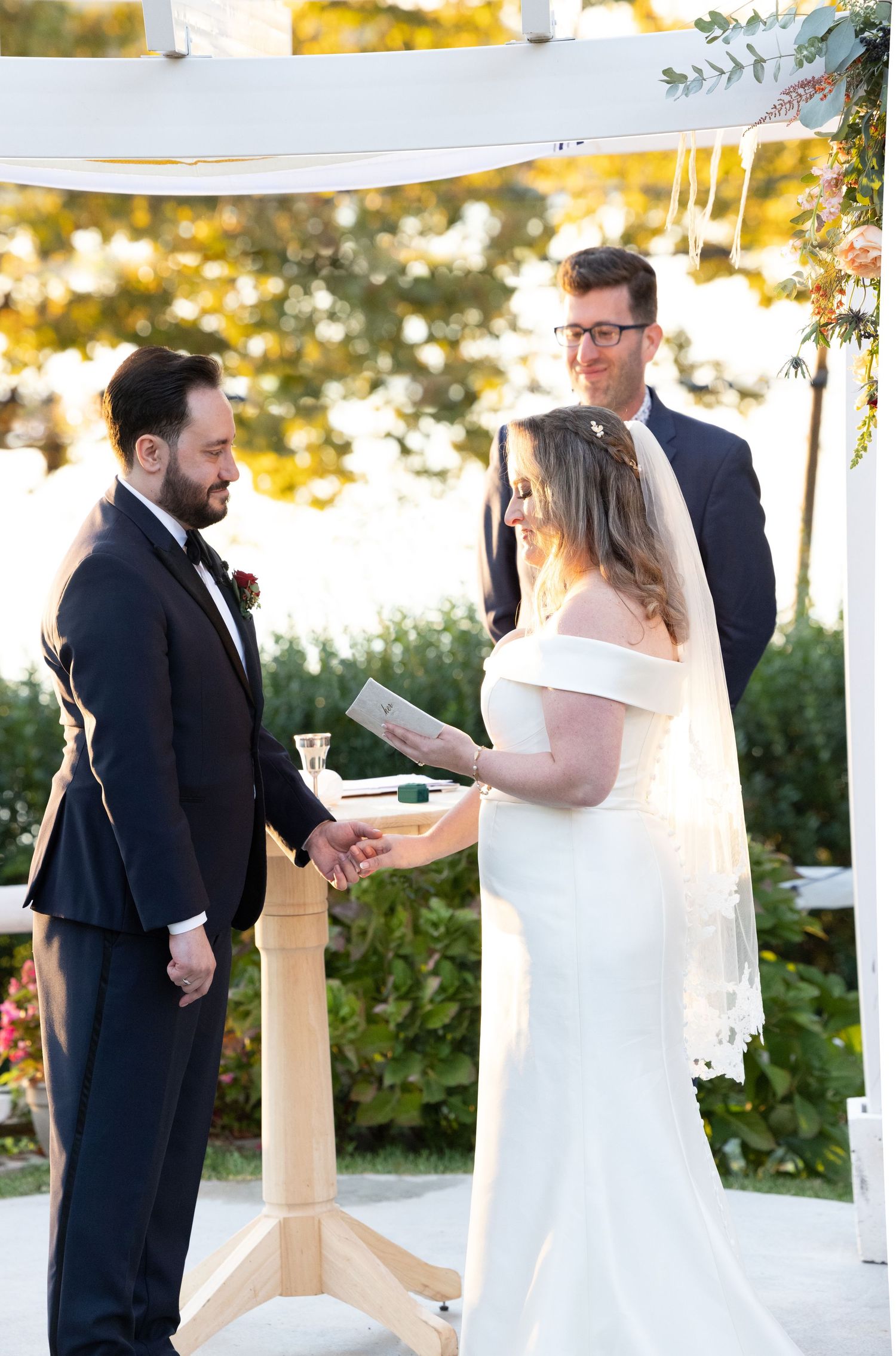 Bride reads vows to groom during outdoor wedding ceremony under floral-decorated white arbor.