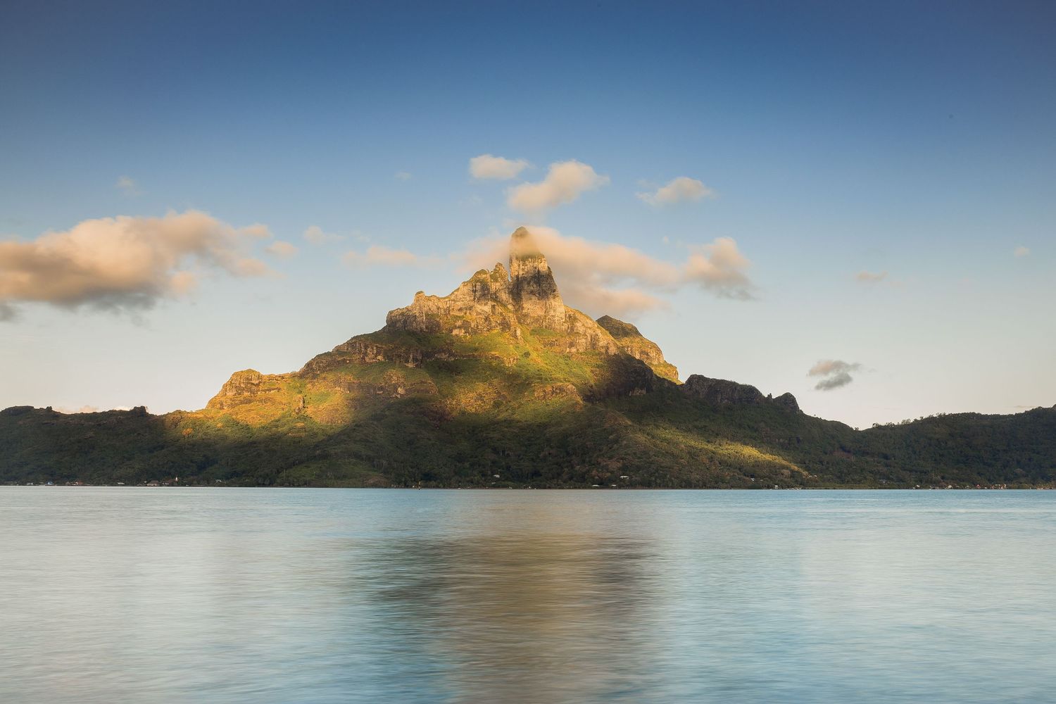 A majestic volcanic mountain peak rises above calm blue lagoon waters under a partly cloudy sky in Bora Bora, French Polynesia.