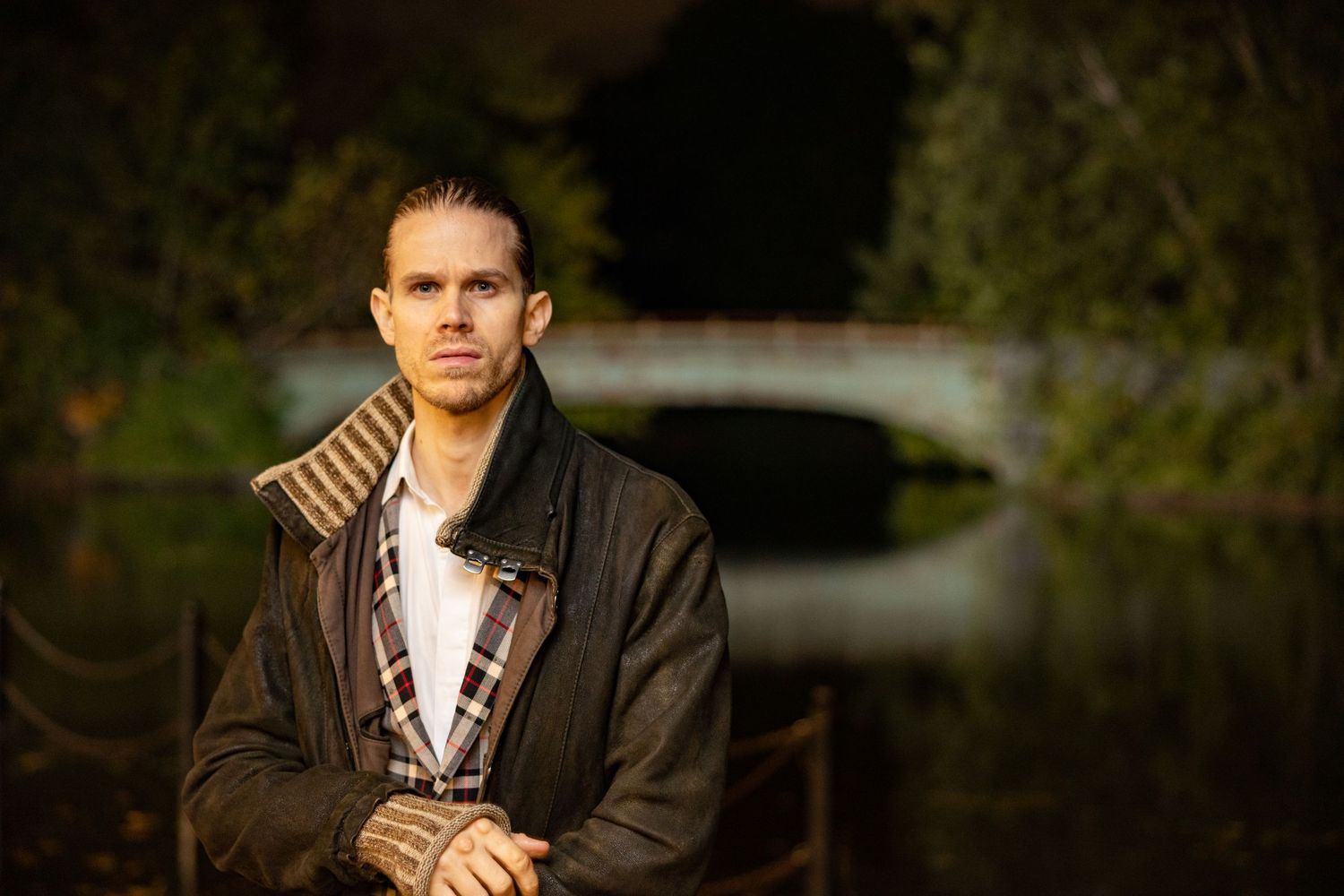 Man in brown leather jacket standing near pond with bridge in background at night.