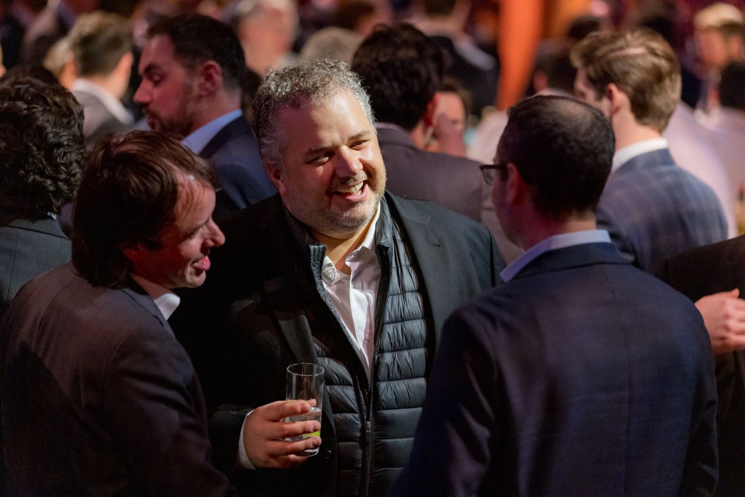 Man in dark jacket smiling and holding drink at crowded networking event.