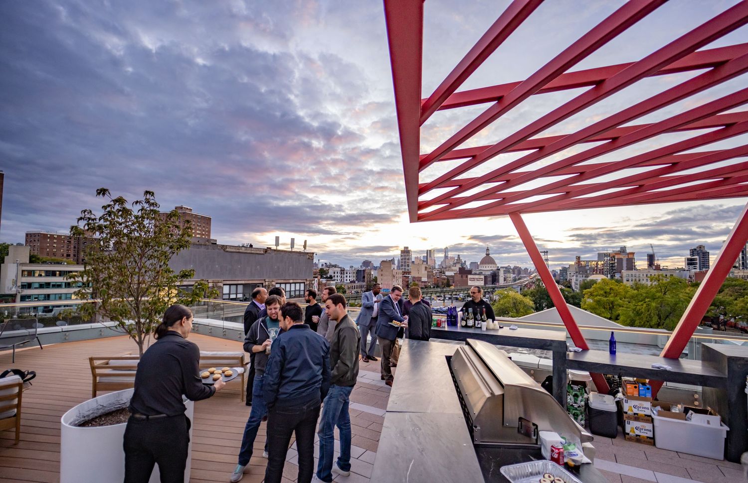 Professionals networking at a rooftop bar event at dusk with city skyline and red architectural structure.