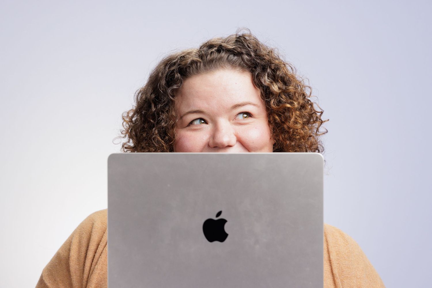 Woman smiling behind an Apple MacBook laptop with curly hair, wearing an orange sweater on white background.