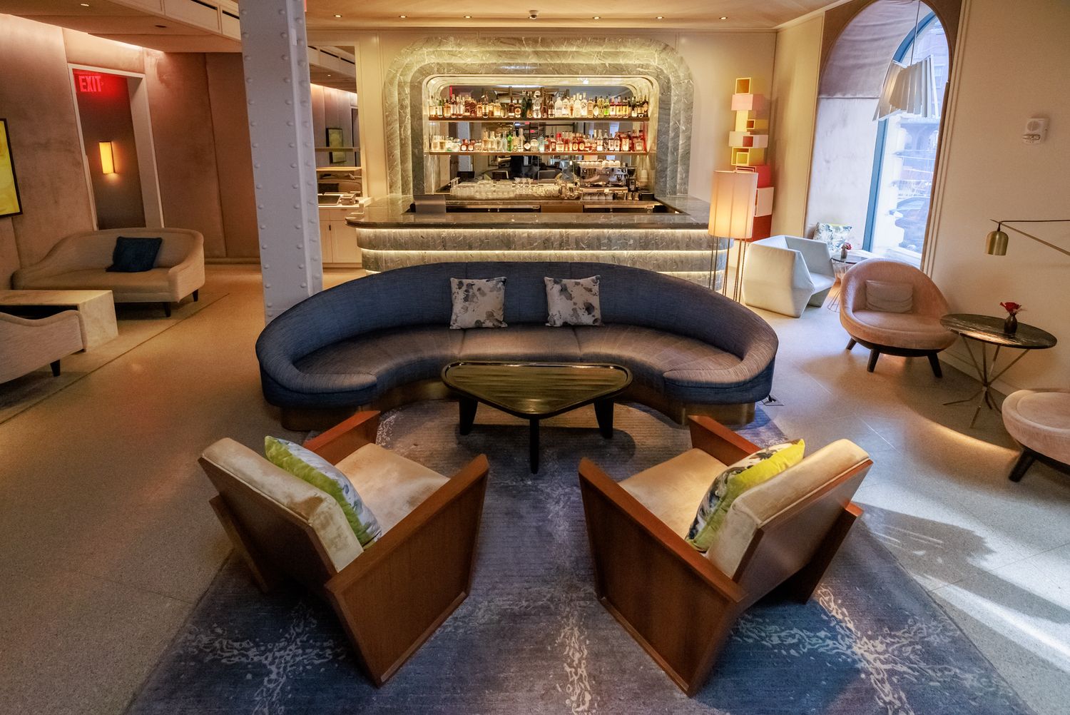Upscale hotel lounge with curved navy sofa, wooden armchairs, and a backlit bar in the background.