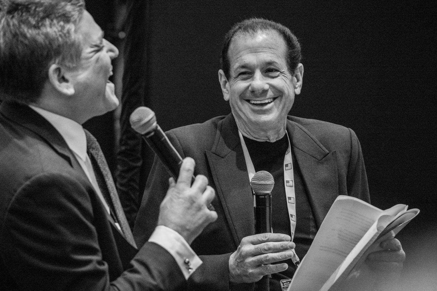 Two men laughing and sharing microphones during a lively black and white conference presentation.