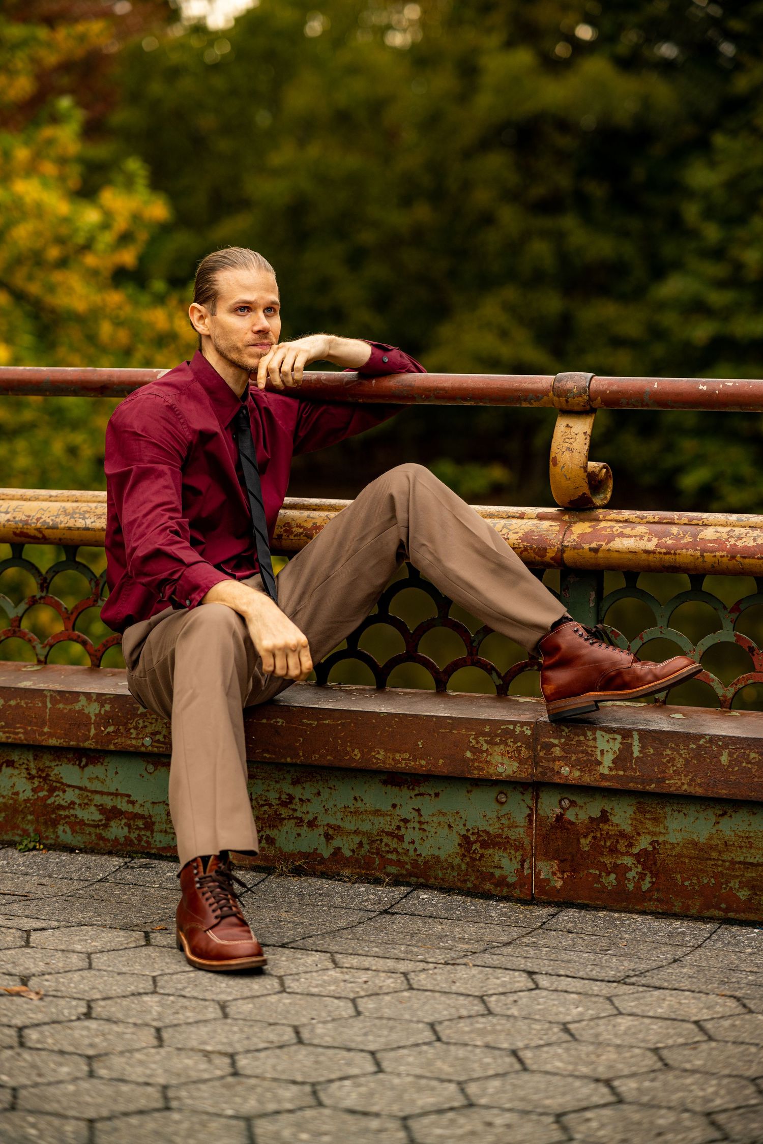 Man in burgundy shirt and brown trousers sitting on rustic bridge railing in green park.