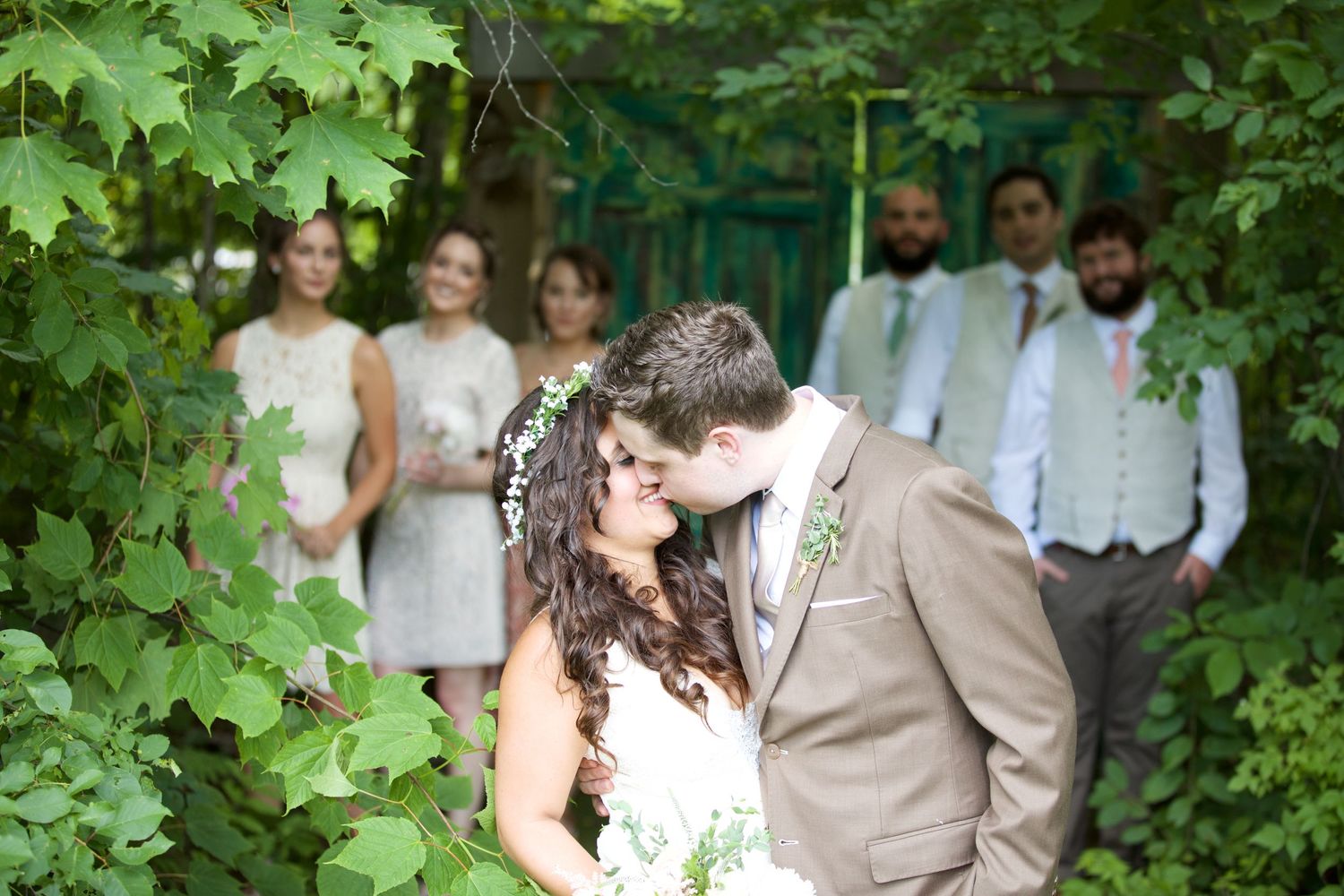 Bride and groom share a romantic moment surrounded by lush green foliage with wedding party in background.