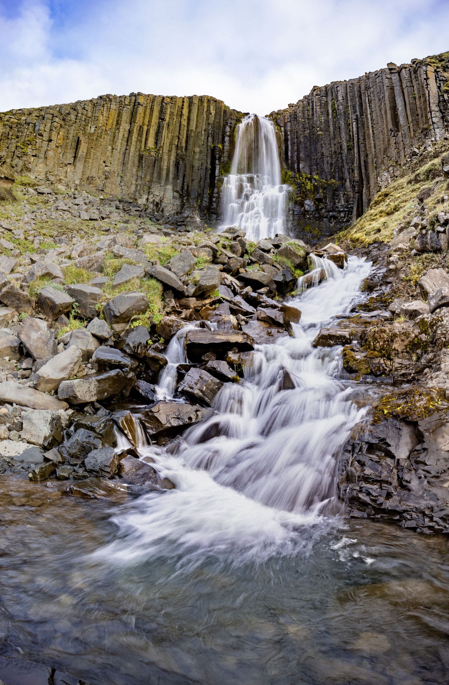 A dramatic waterfall flows down geometric basalt column formations onto rocky terrain in an Icelandic landscape.