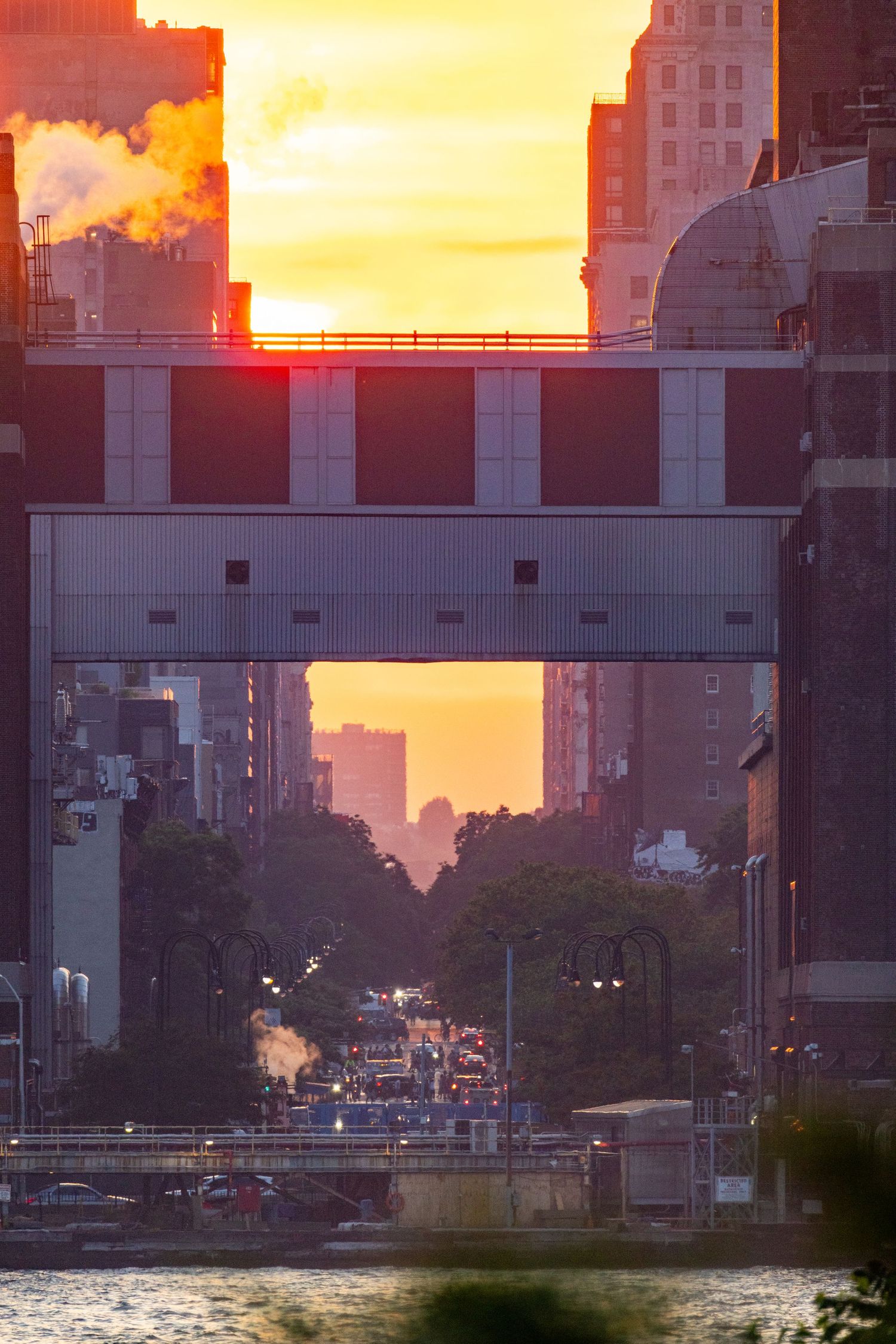 Golden sunset Manhattanhenge phenomenon illuminating busy New York City streets viewed through an elevated bridge.