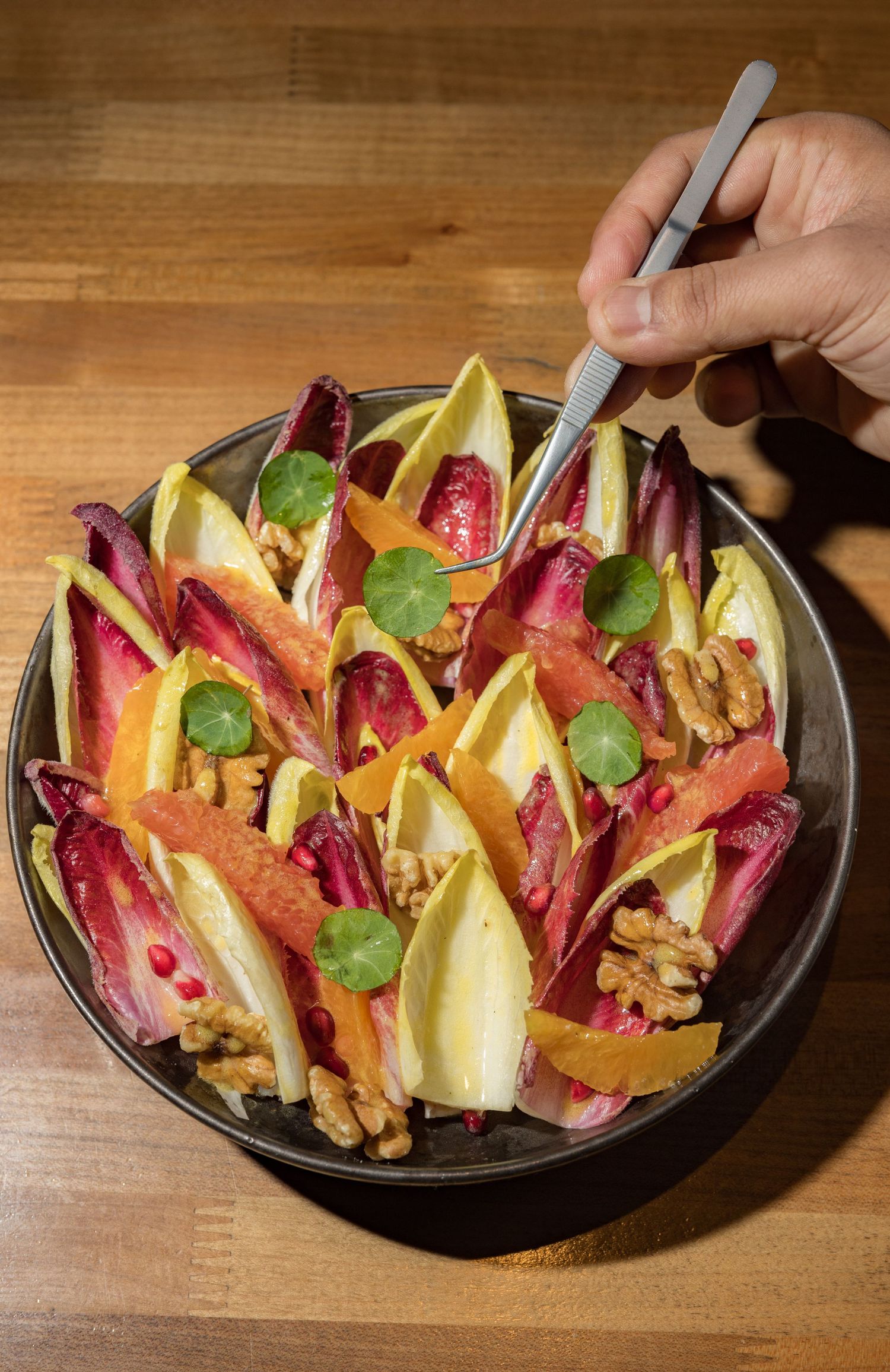 Colorful endive salad with citrus segments, walnuts, pomegranate seeds, and fresh herbs in a dark bowl.