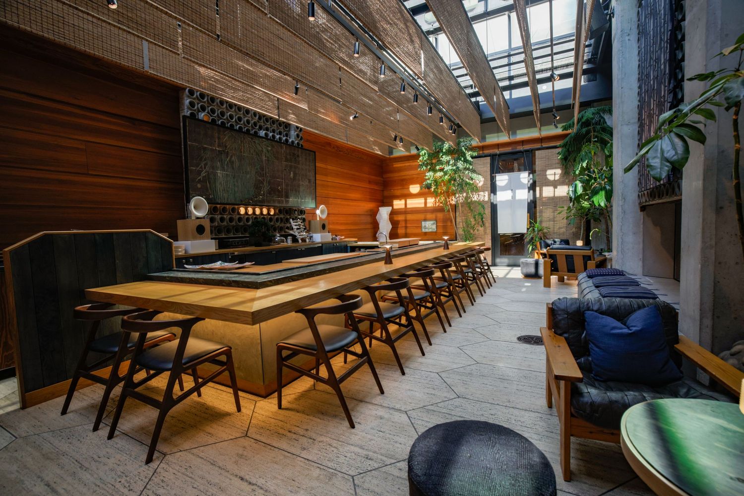 Modern restaurant interior with long wooden bar, black stools, skylights, and lush green plants.