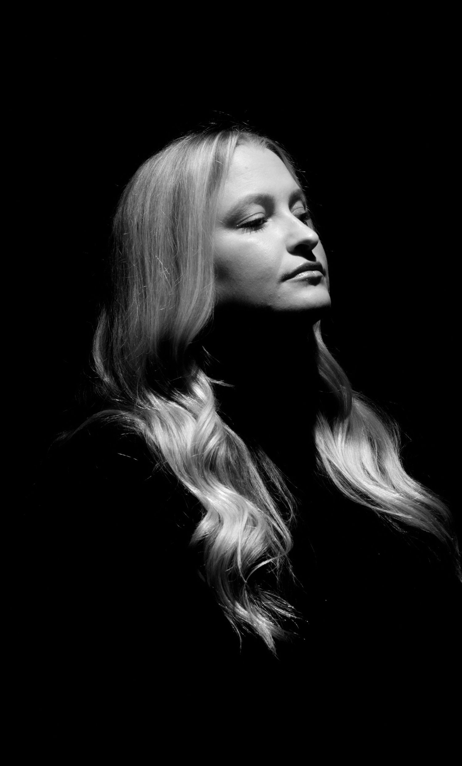 Black and white portrait of a blonde woman with closed eyes against a dark background.